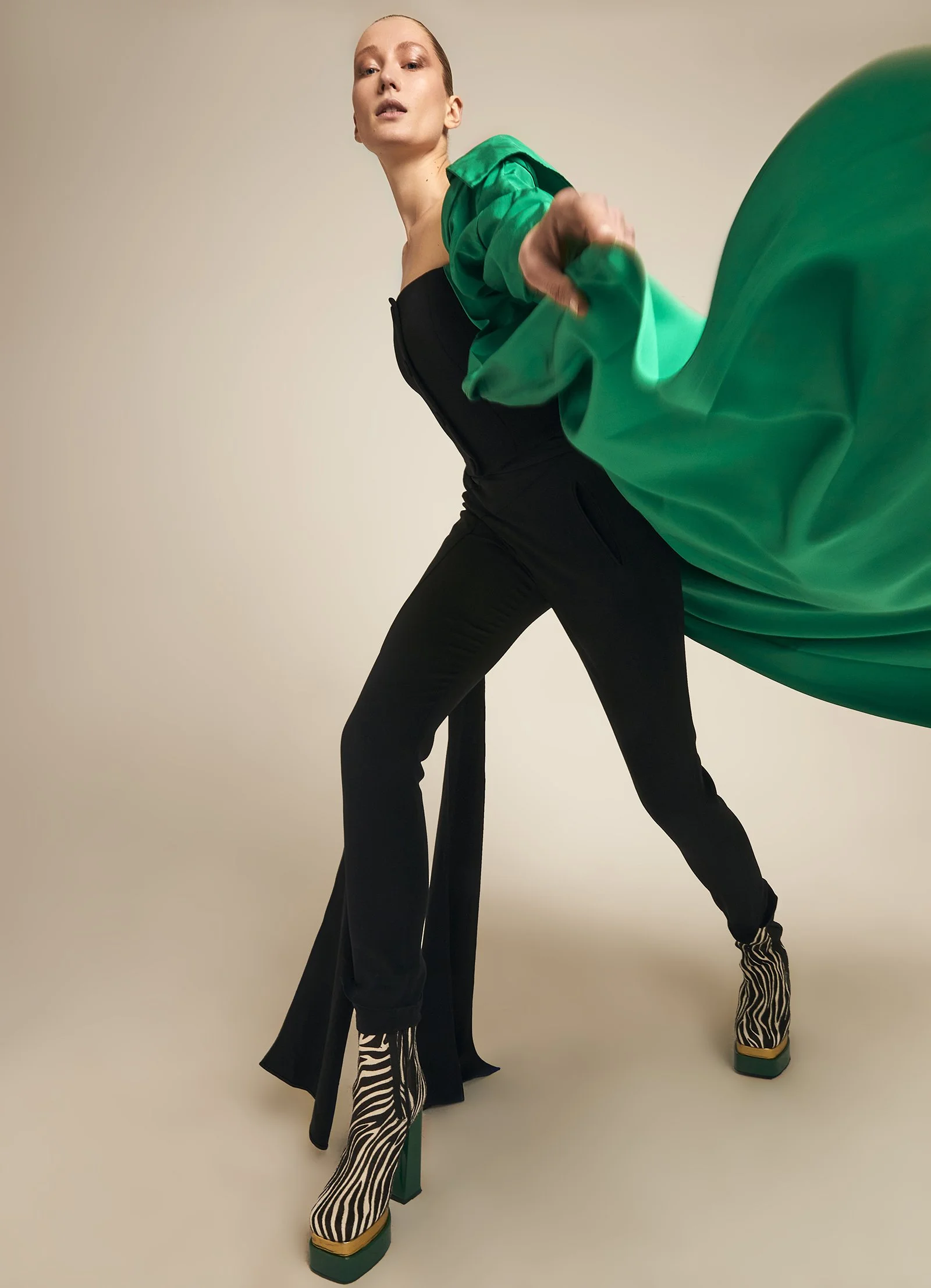 Fashion model in black outfit with dramatic green cape, wearing zebra-patterned platform boots, beige background.