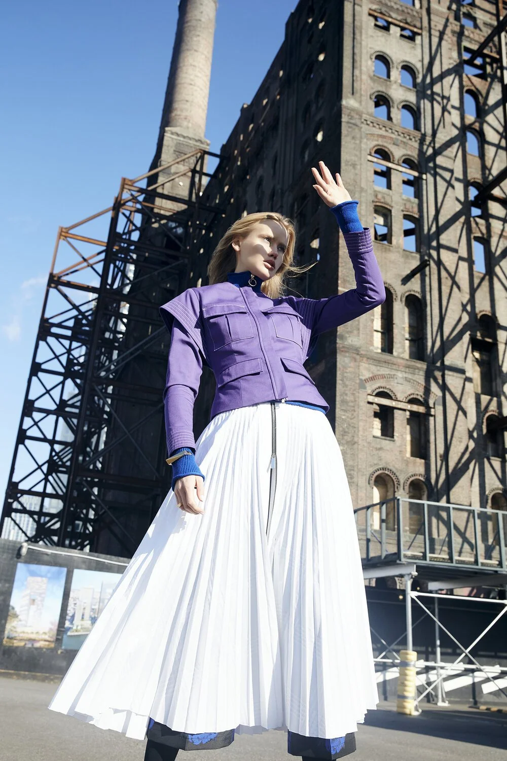 Woman in purple jacket and white pleated skirt standing in front of an industrial building, sunny day.