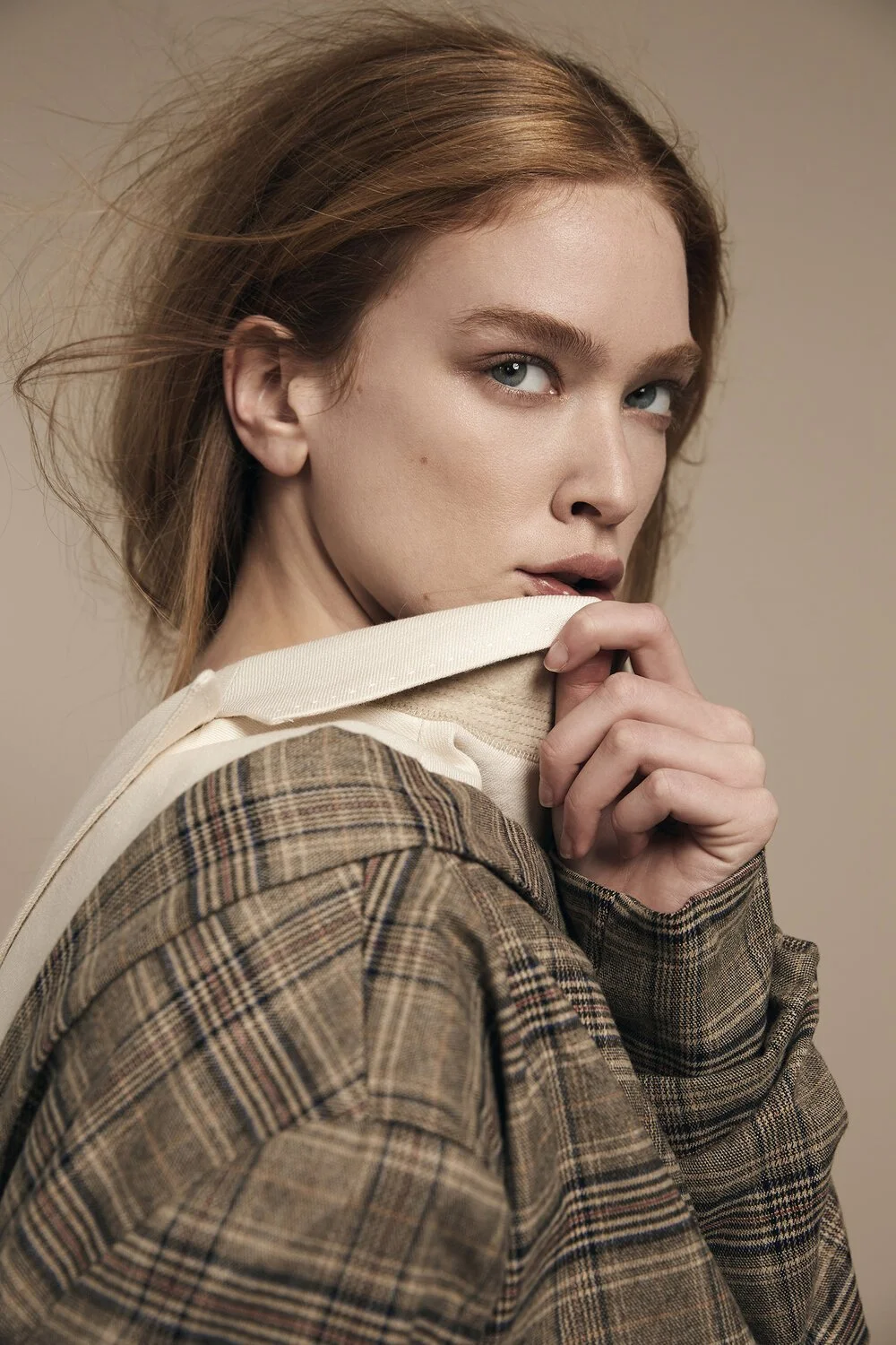 Person with light brown hair wearing a plaid jacket and beige shirt, posing thoughtfully with a hand near the mouth.