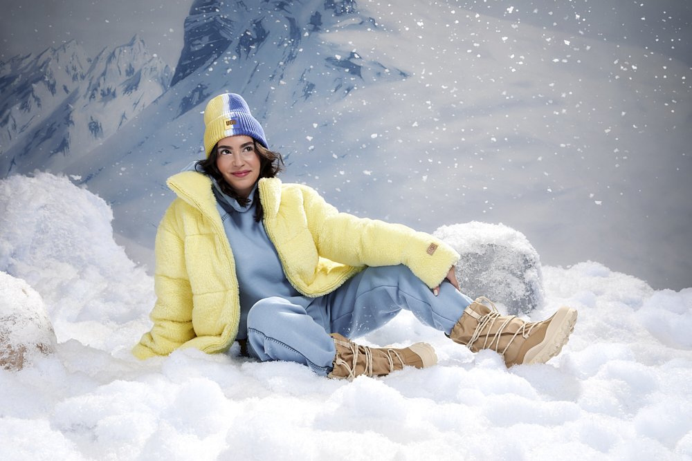 Model wearing a yellow puffer jacket, blue sweatsuit, and tan Ugg winter boots in a snowy studio set, commercial fashion photography by Adrianna Favero, NYC-based photographer and director, for the Ugg x Travel + Leisure campaign.
