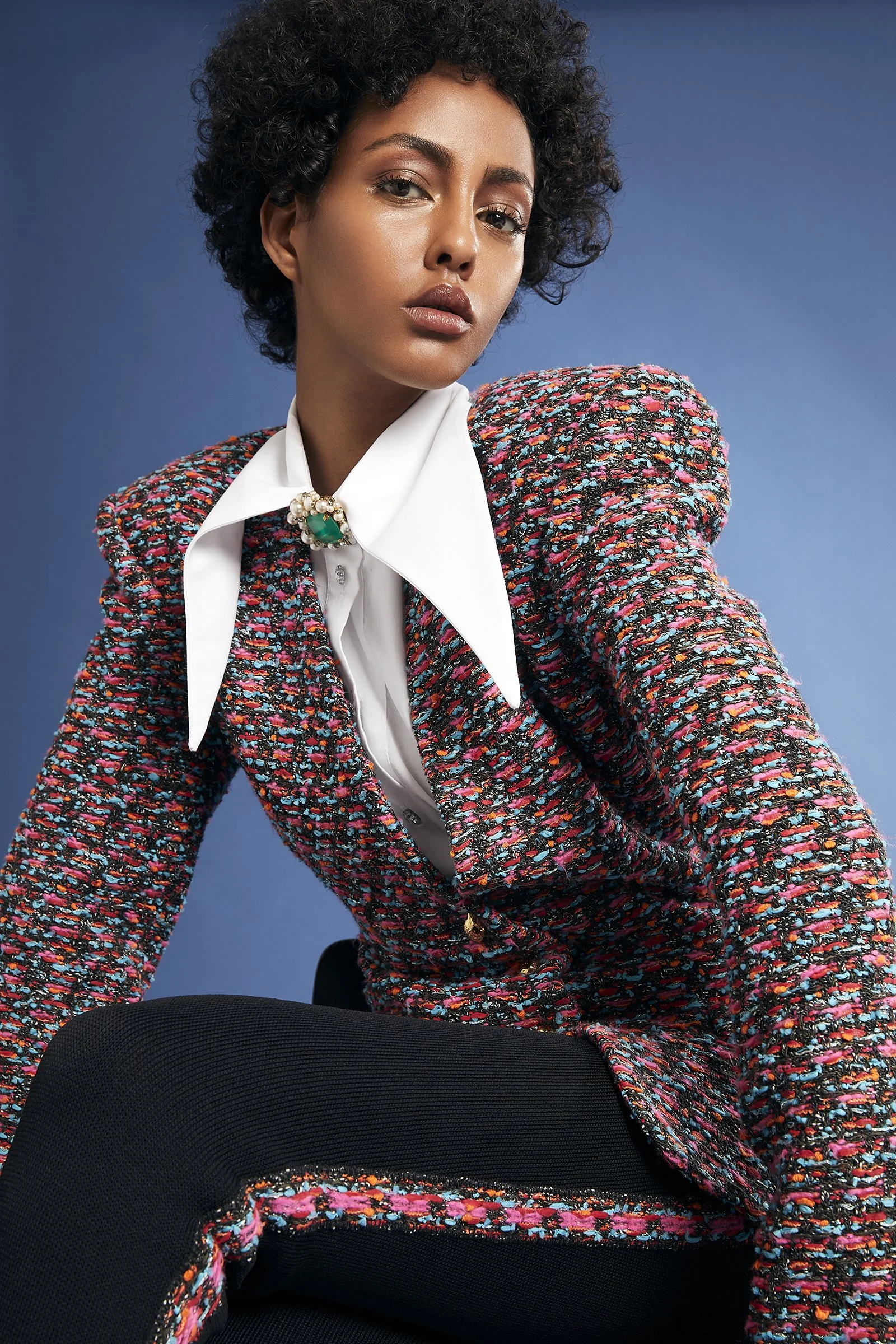 Fashion model wearing a colorful tweed jacket and black pants, with a white shirt and statement collar, posed against a blue background.
