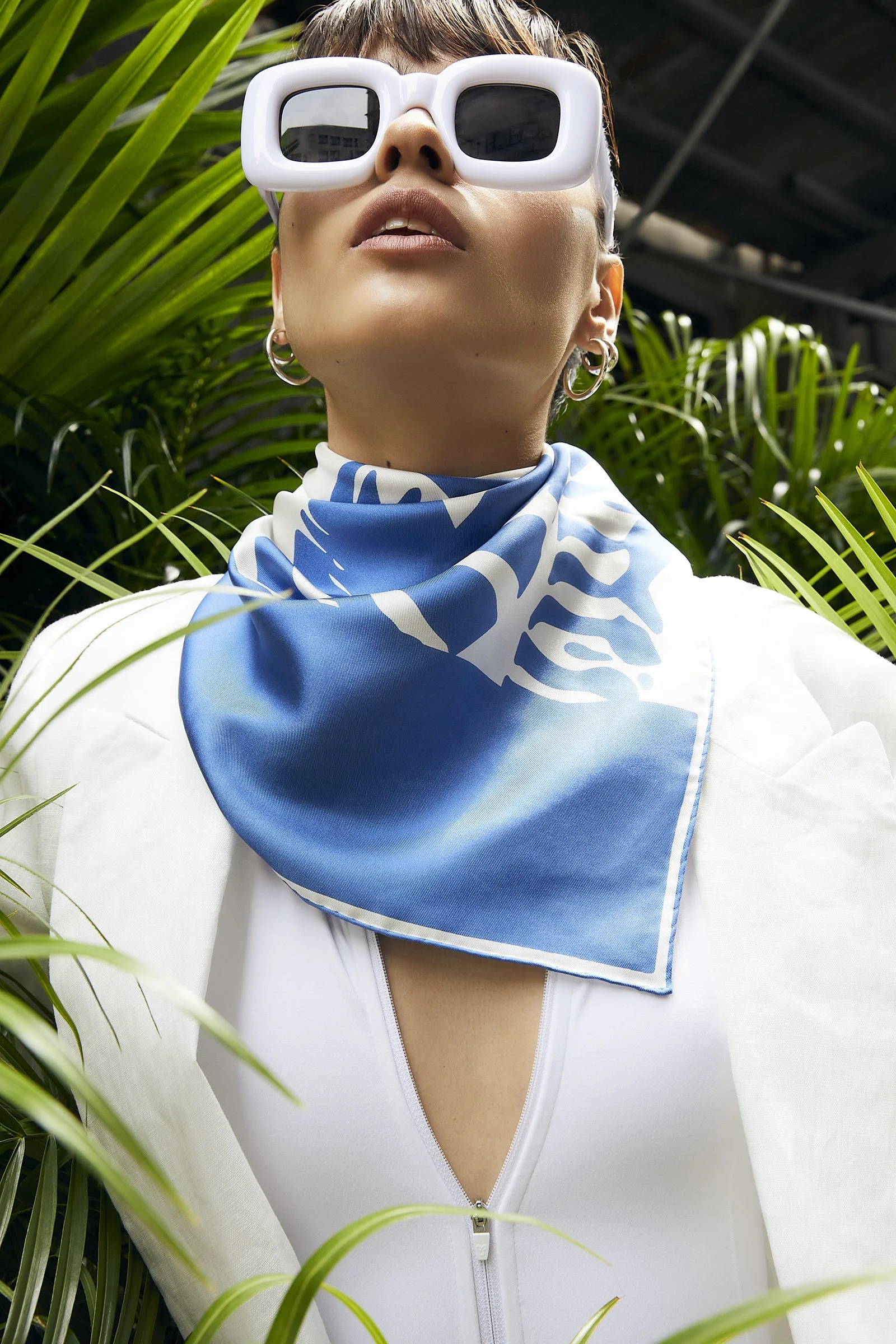 Person wearing oversized white sunglasses, large hoop earrings, a blue and white scarf, white zippered top, surrounded by green plants.