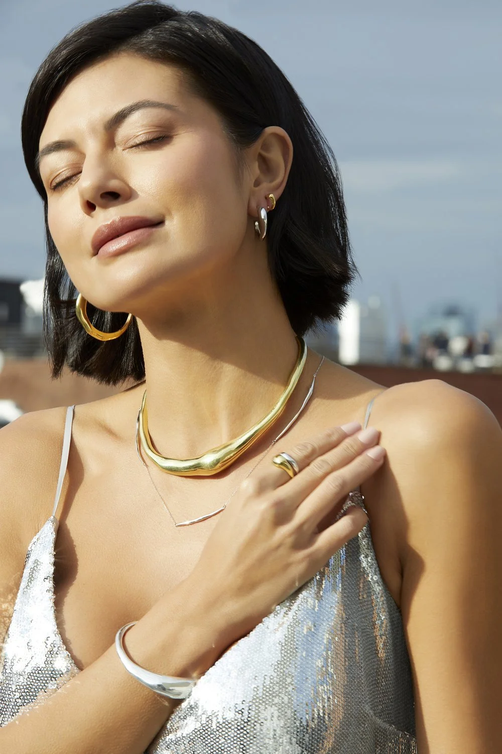 Woman with closed eyes, wearing gold and silver jewelry, including earrings, necklaces, bracelet, and ring, in a reflective silver top, outdoors.