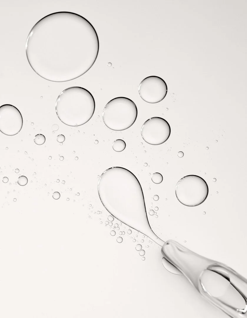 Close-up of clear gel serum droplets with a dropper applicator on a white background, by Adrianna Favero.