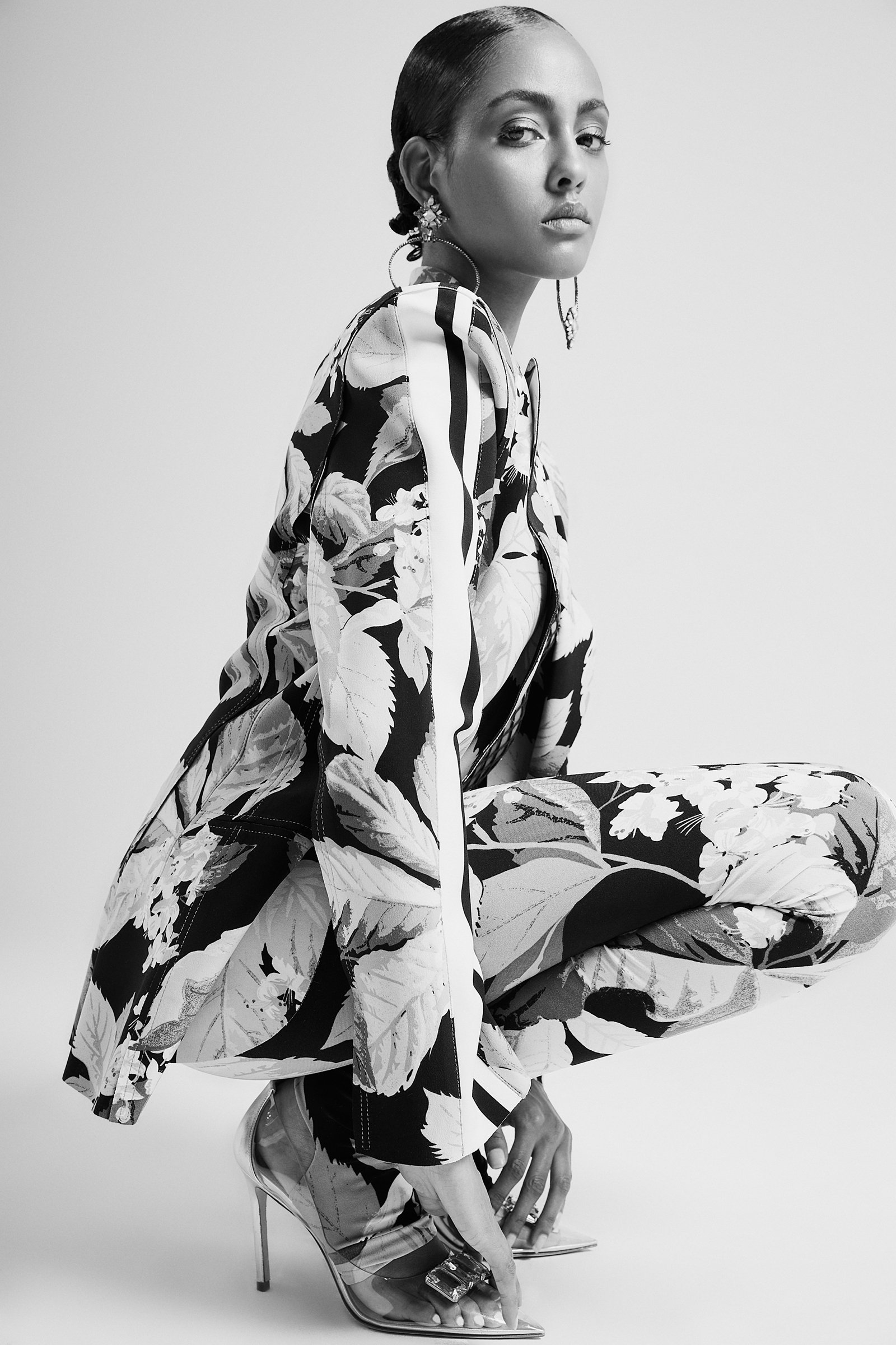 Black and white portrait of a fashion model wearing a floral patterned suit and high heels, kneeling with a confident expression.