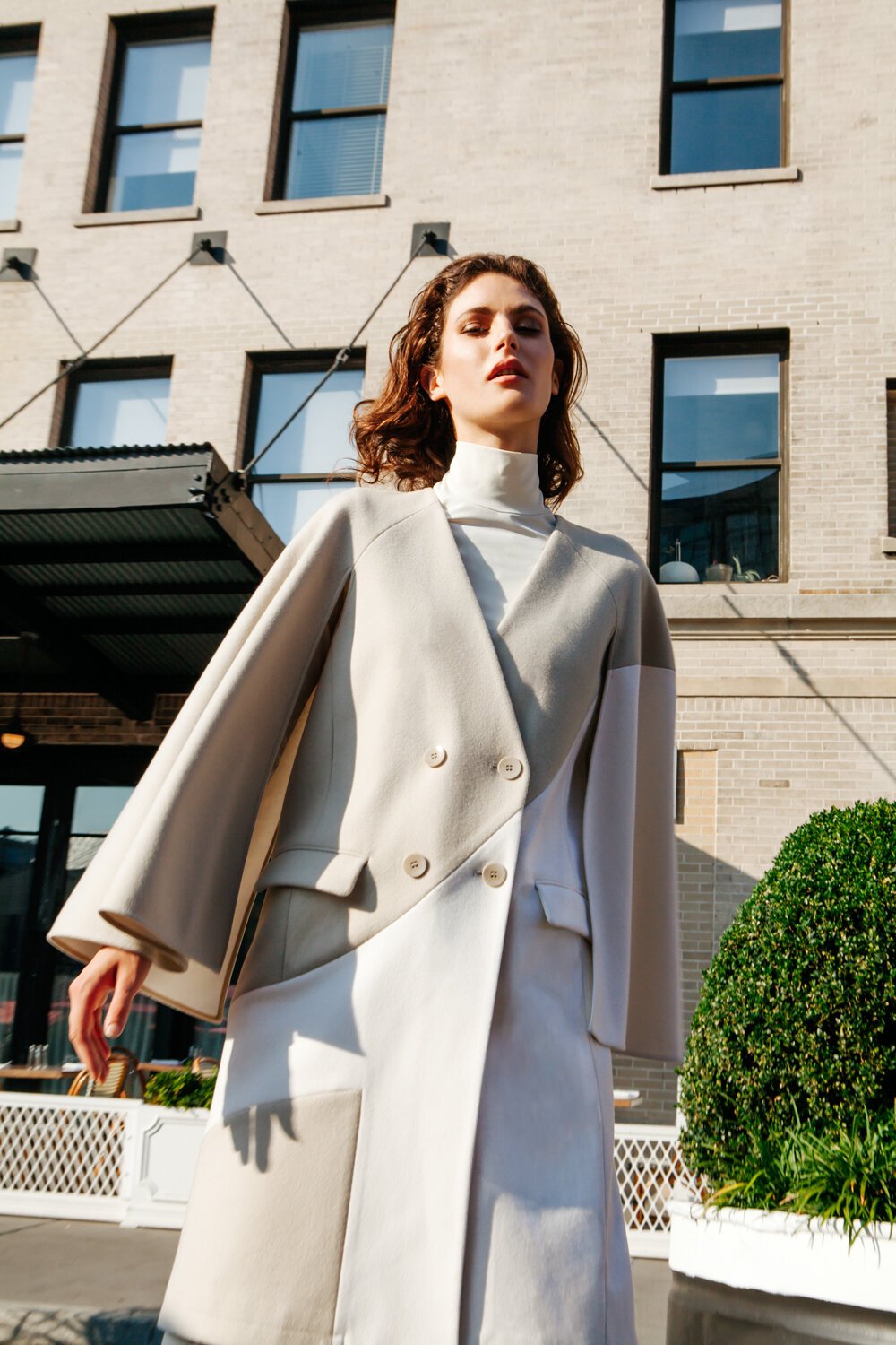 Fashion model wearing a double-breasted coat and turtleneck, posing outdoors in an urban setting, with a brick building in the background.