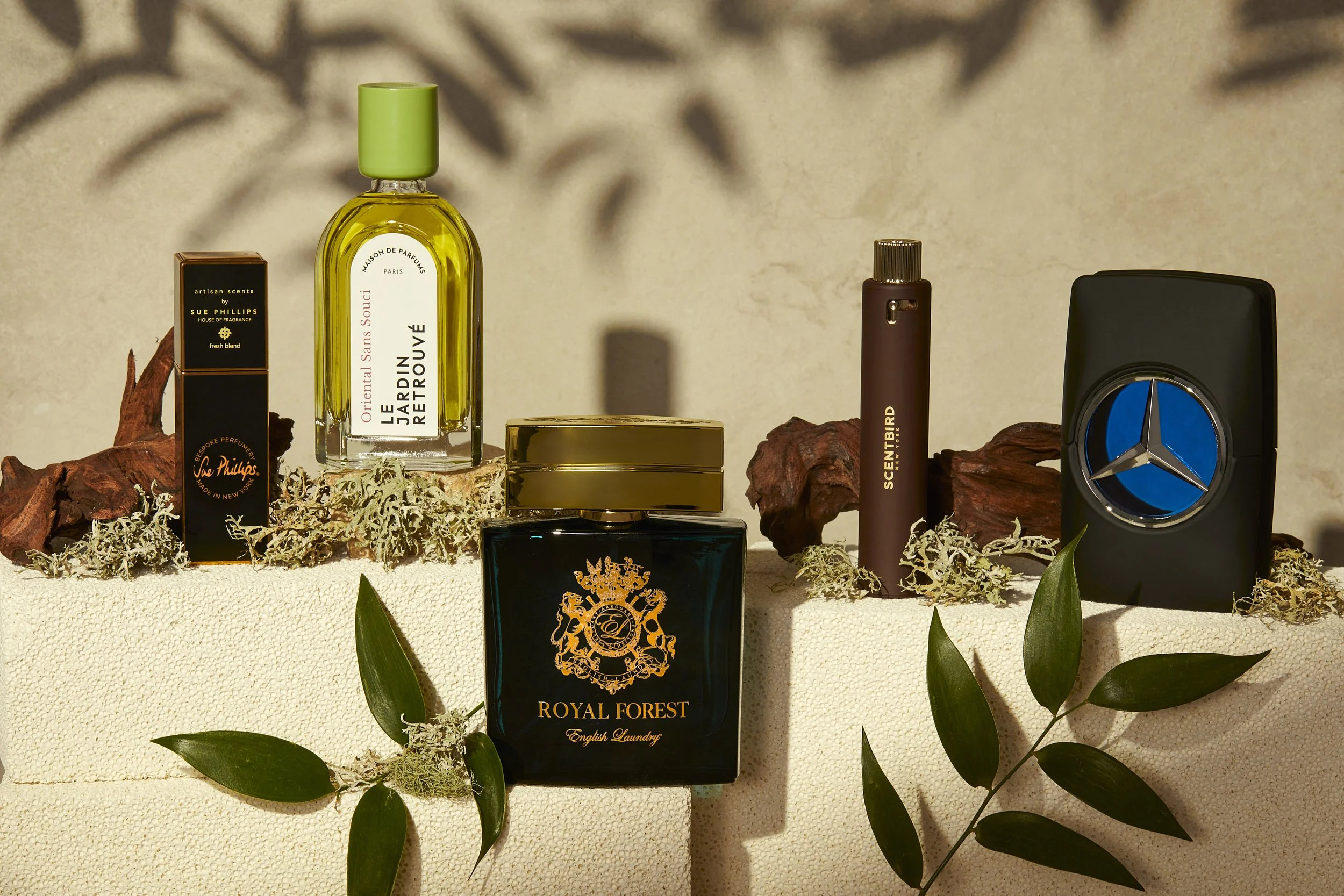 Arrangement of various perfume bottles and boxes on a textured surface with green leaves and decorative moss, including brands like Sue Phillips, Le Jardin Retrouvé, Scentbird, Royal Forest, and a blue emblem bottle.