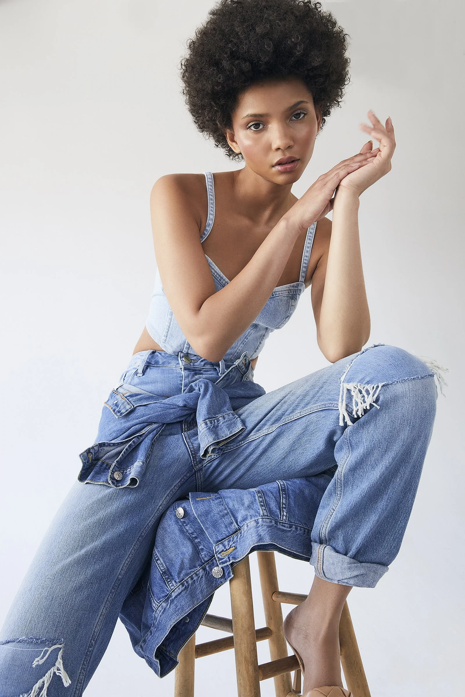 Person in denim outfit sitting on a wooden stool, with short curly hair and casual pose.
