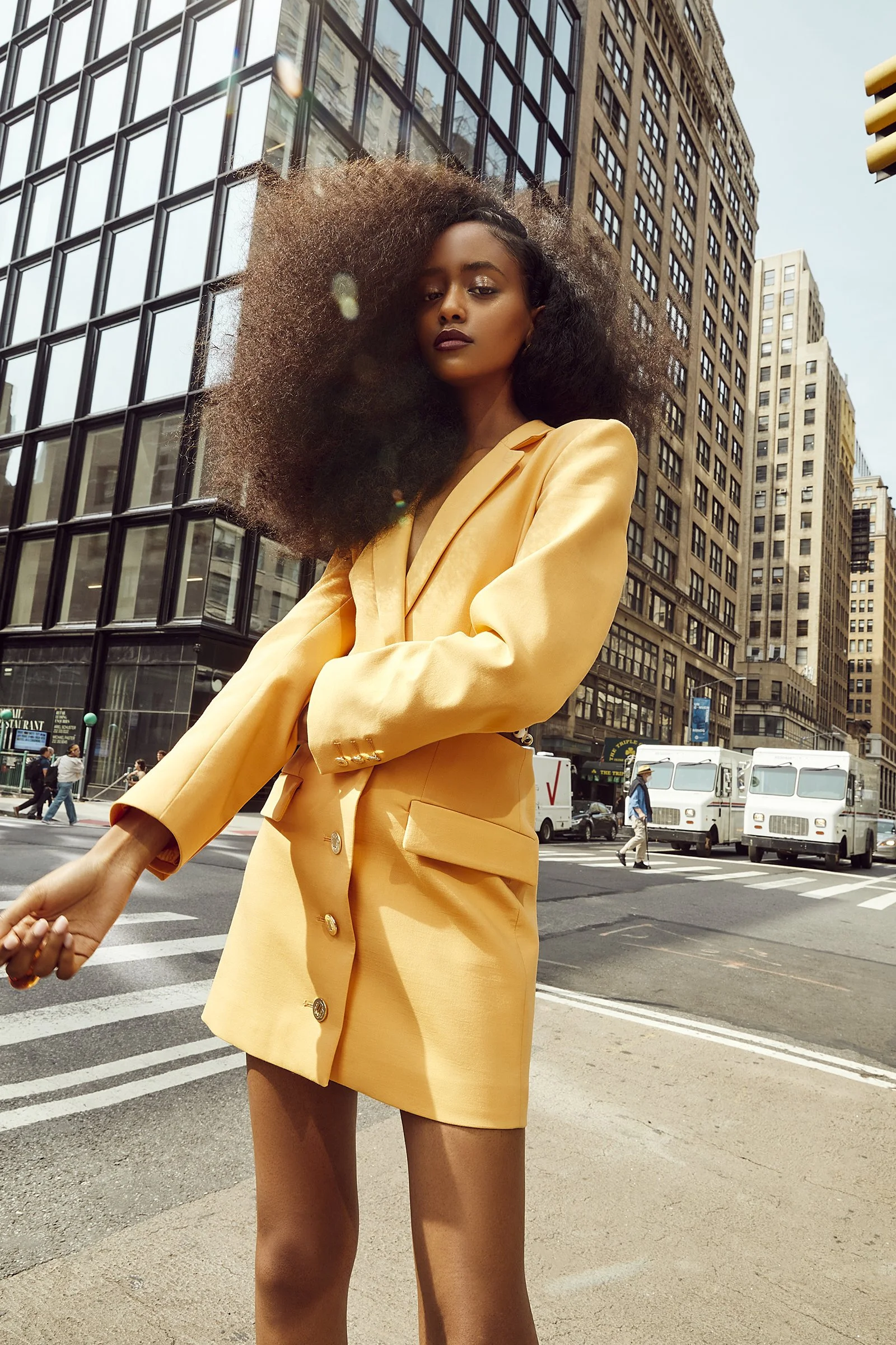 Fashionable woman in a yellow blazer dress with voluminous hair standing on a city street corner.