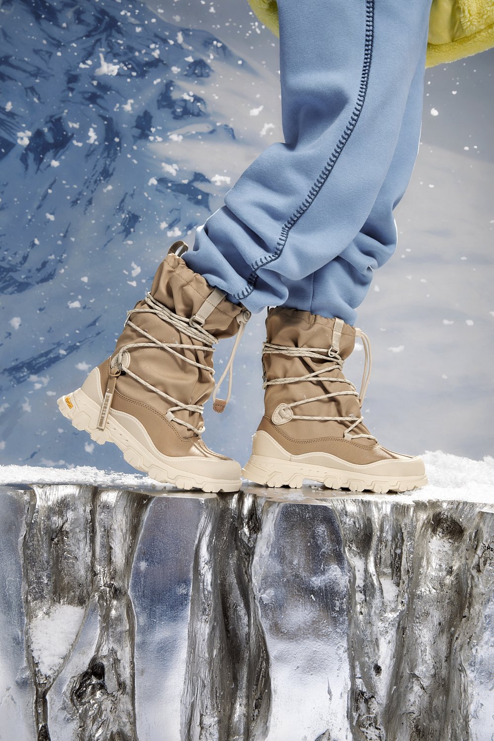 Person wearing tan hiking boots with thick soles, standing on an icy surface; background shows a snowy, mountainous landscape.