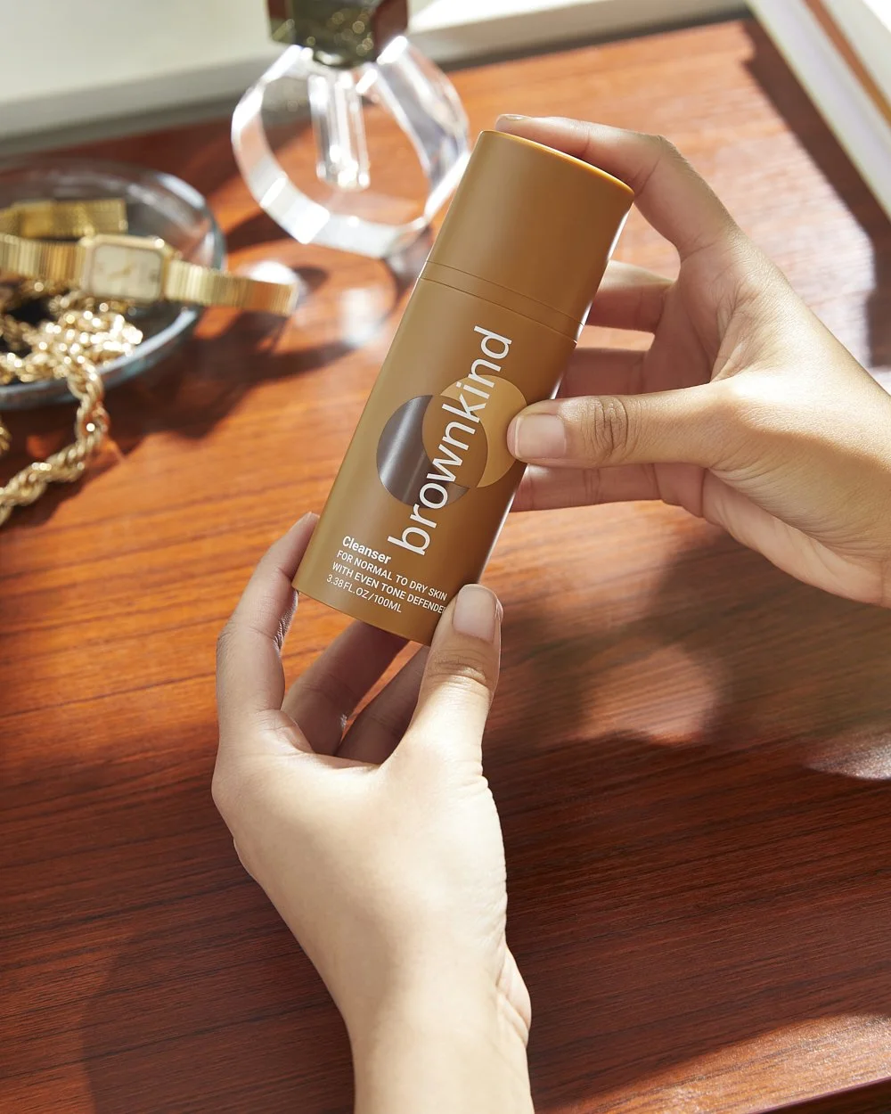 Brownkind lifestyle still life: hand holding the Brownkind serum bottle above a warm-toned styled surface with jewelry, photographed by Adrianna Favero.