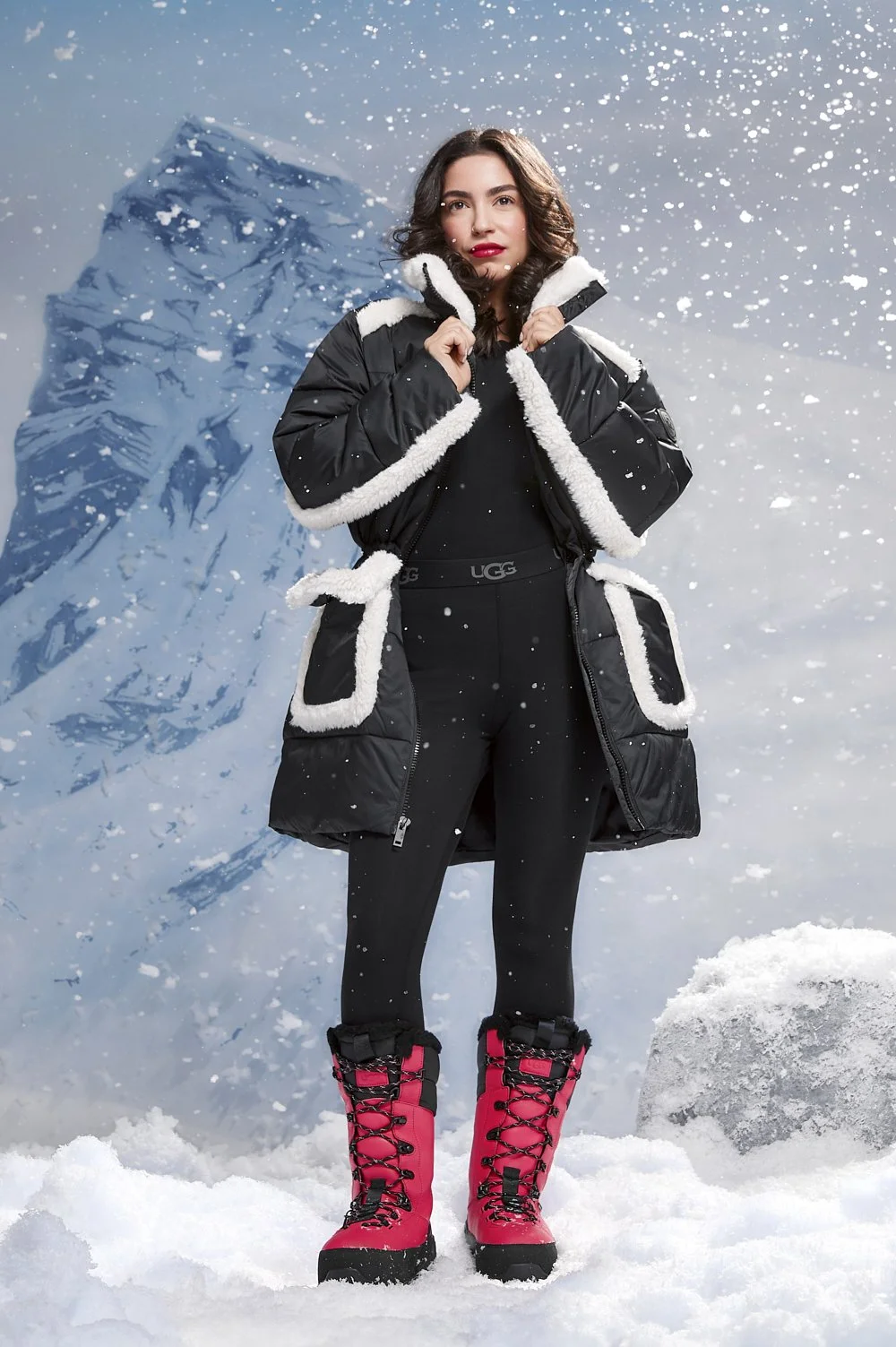 Person wearing winter clothing in snowy mountain setting, featuring a black coat with white fur trim and red snow boots.