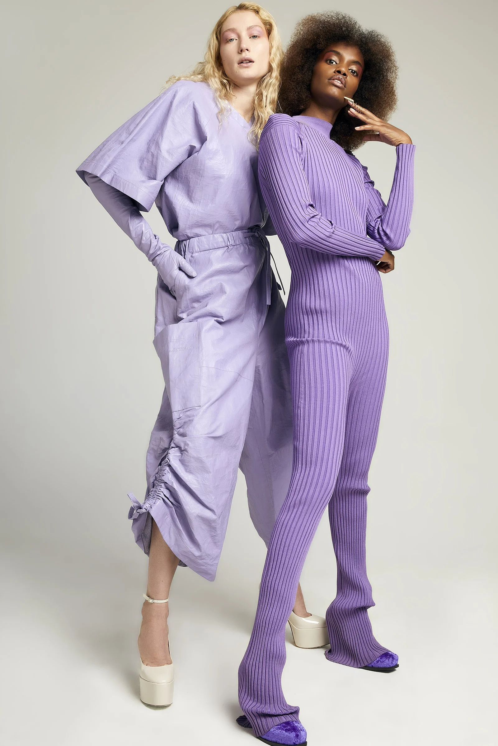Two models wearing purple outfits, one in a shiny dress with wide sleeves and the other in a ribbed jumpsuit, both wearing platform heels.