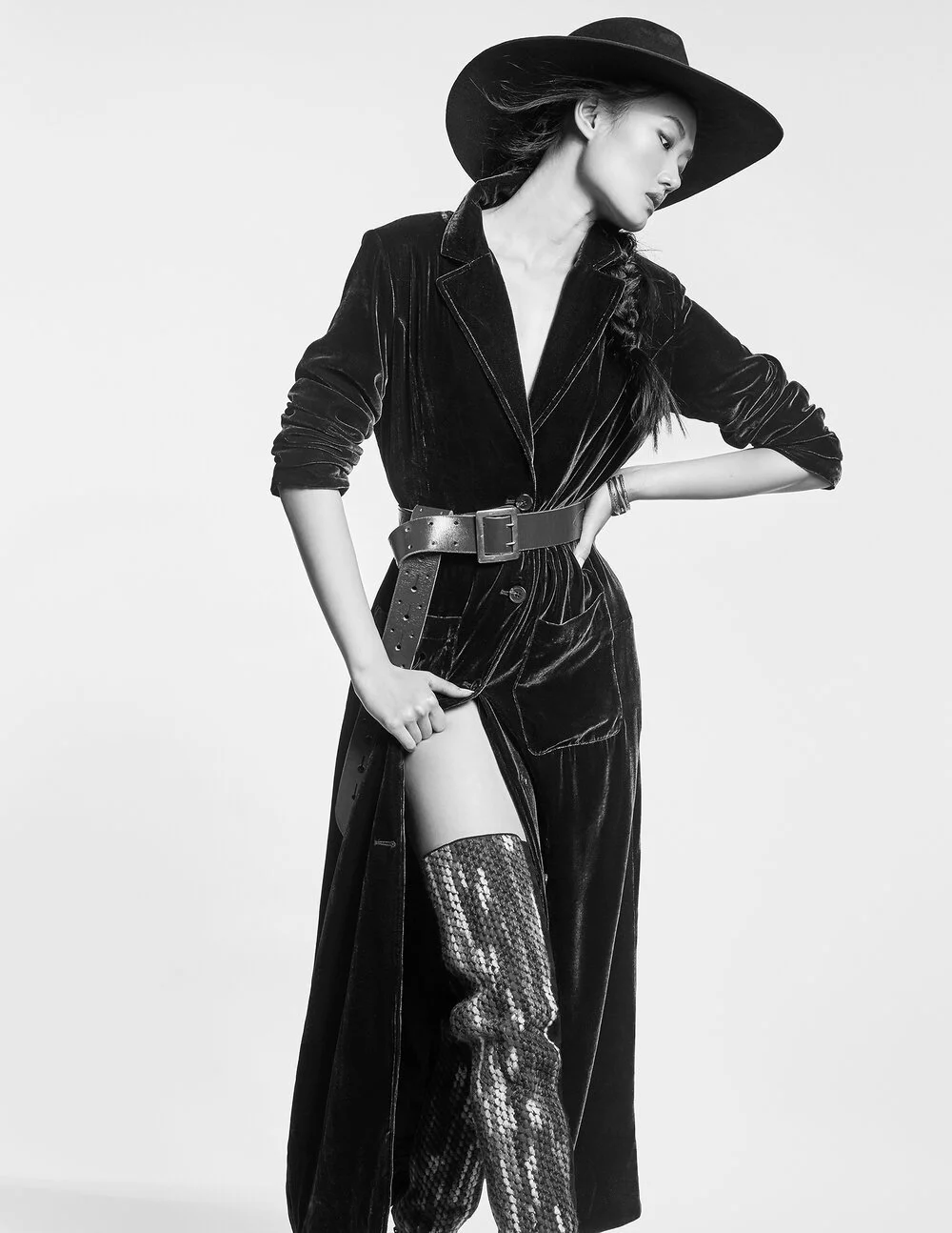 Fashion model wearing a long velvet coat, wide-brimmed hat, and sequin thigh-high boots, posing in a black and white image.