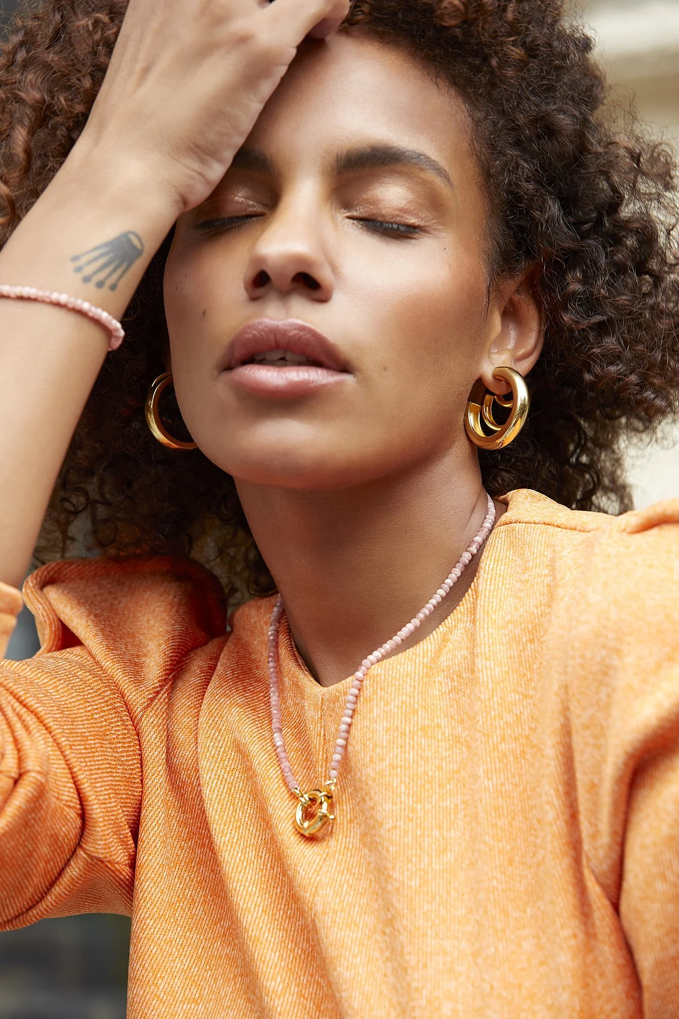 Person wearing an orange top and gold hoop earrings, with one hand on their forehead, eyes closed, and a pink bead necklace.