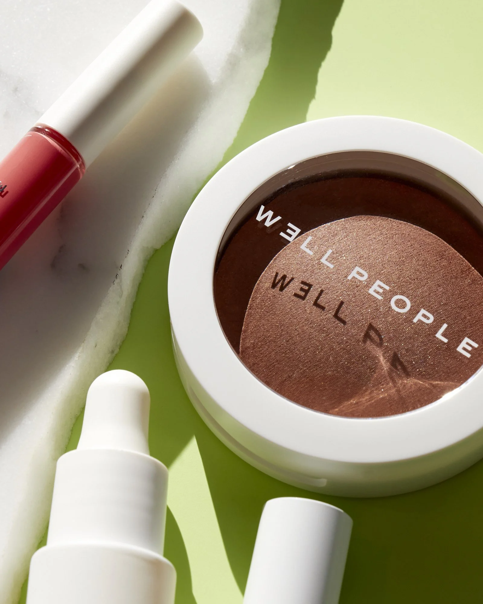 Cosmetic products including a compact of bronzer, a pink lip gloss, and a white bottle with a dropper on a green and marble backdrop.