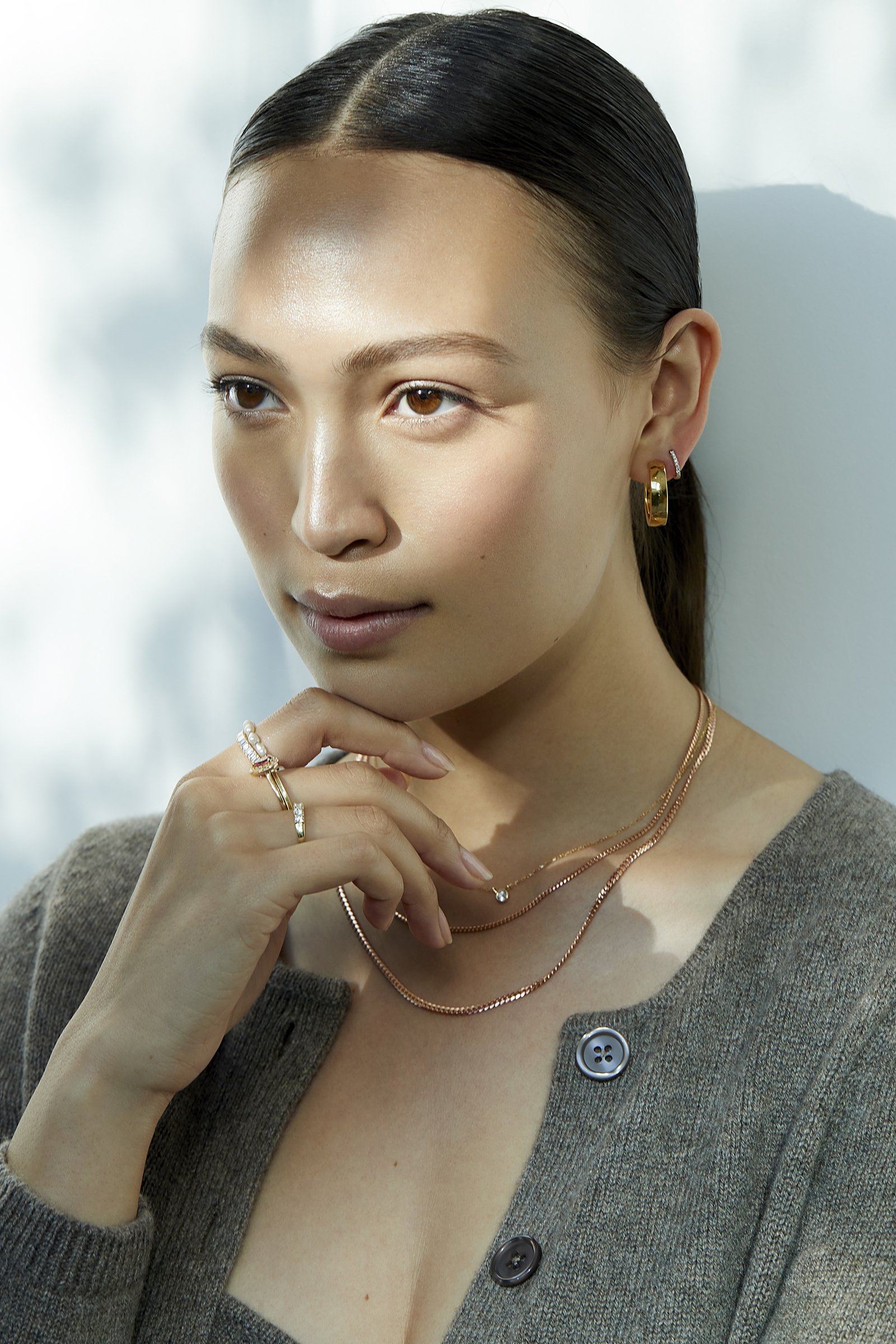 Model resting her fingers lightly on her jaw, wearing a layered gold necklace and a gray cardigan, soft natural lighting with a blurred background, for an Aurate New York jewelry editorial.
