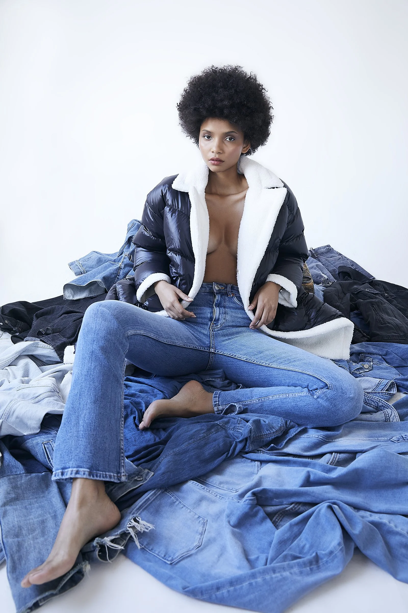 Person wearing a black puffer jacket with white lining and blue jeans, sitting on a pile of denim clothing, against a plain background.
