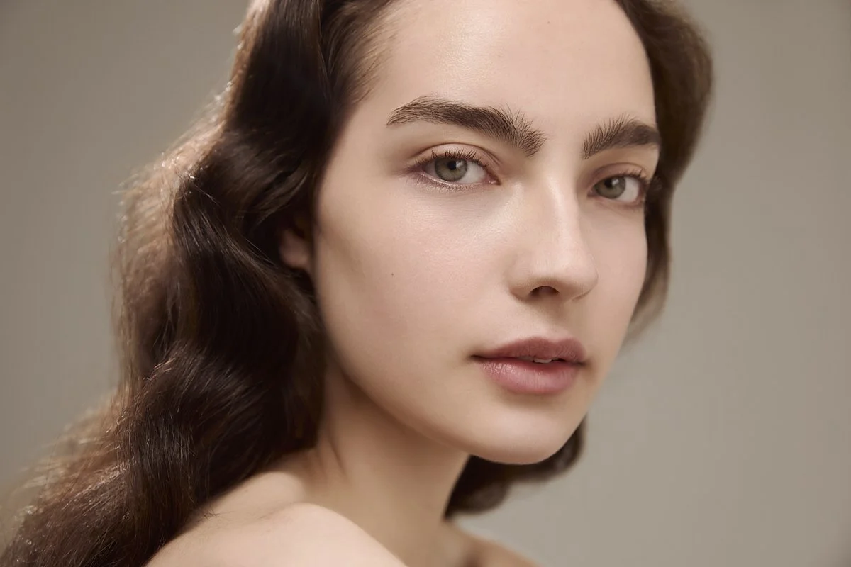 Beauty portrait of a model with dark wavy hair and natural makeup against a warm neutral background, showcasing radiant, healthy skin for a beauty editorial campaign by Adrianna Favero, NYC beauty photographer and director.