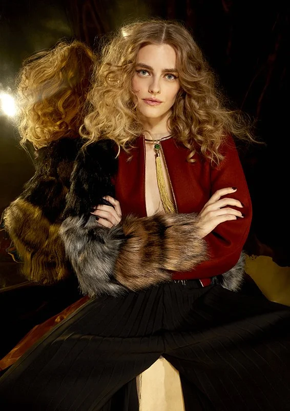FAJO Magazine holiday fashion editorial: close portrait of a model with voluminous curly hair in a black and burgundy outfit, holding a fur stole, photographed by Adrianna Favero.
