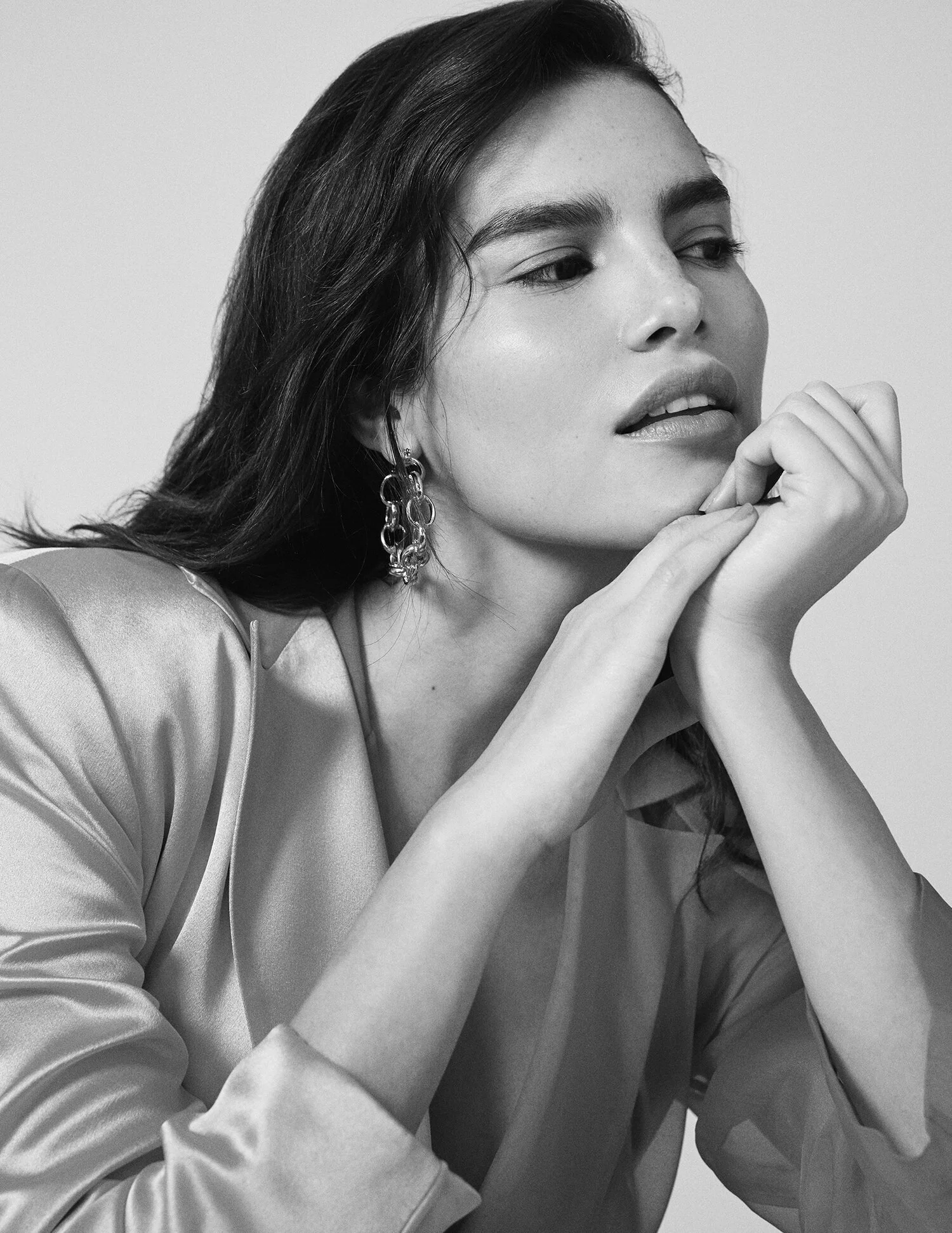 Black and white portrait of a woman with long hair wearing statement earrings and a silk blouse, resting her chin on her hands, looking thoughtfully into the distance.