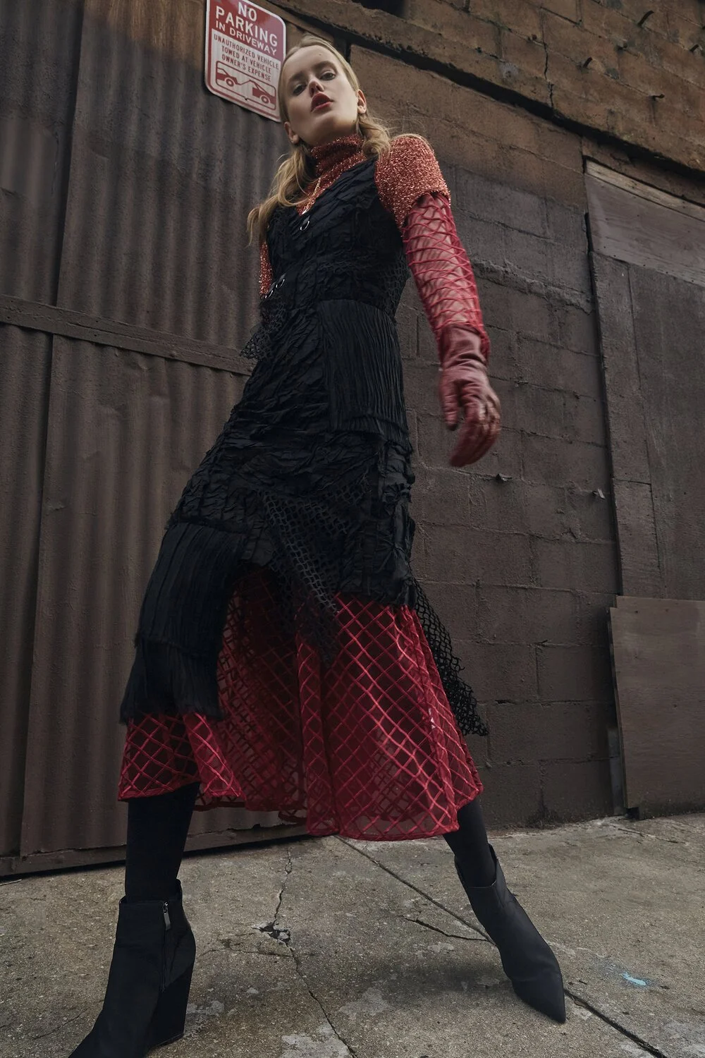 Fashion model wearing a layered red and black dress, posing in an urban alleyway under a 'No Parking' sign.