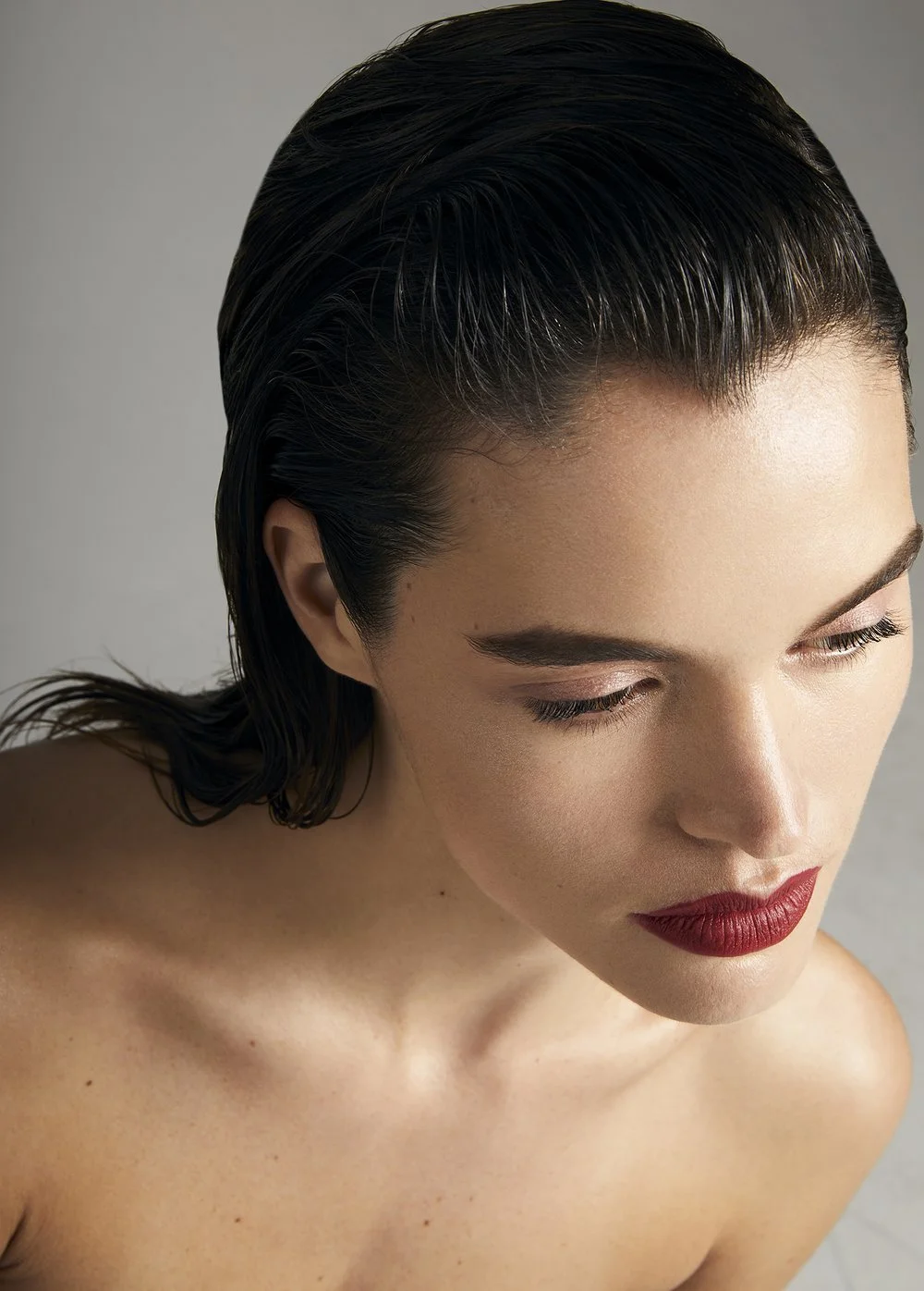 Close-up beauty portrait of a model with sleek wet-look dark hair and bold red lipstick against a neutral gray backdrop, photographed by NYC beauty photographer Adrianna Favero for Gritty Pretty.