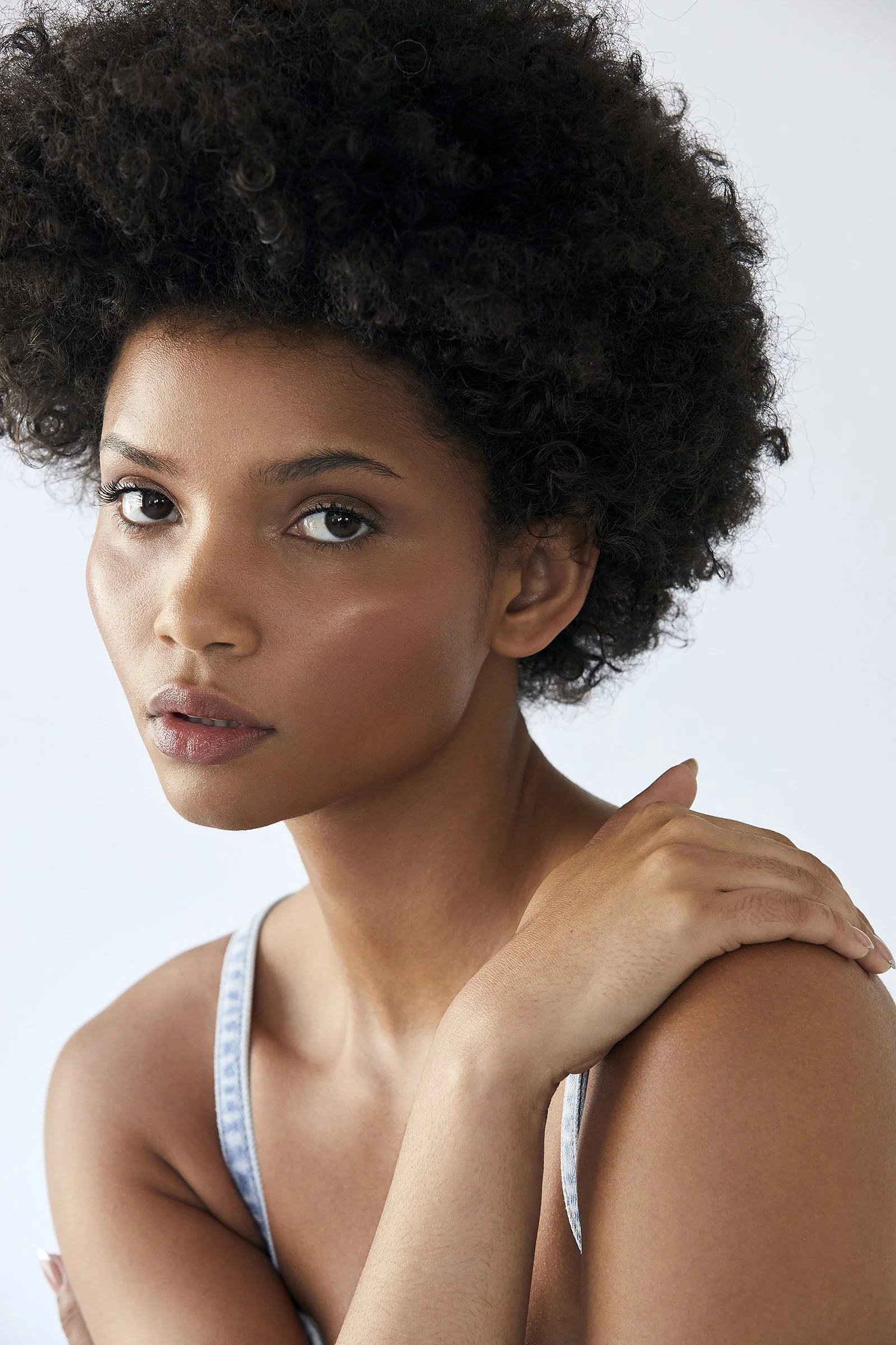 A woman with natural curly hair looking at the camera, resting her hand on her shoulder. She has a neutral expression and is wearing a sleeveless top.