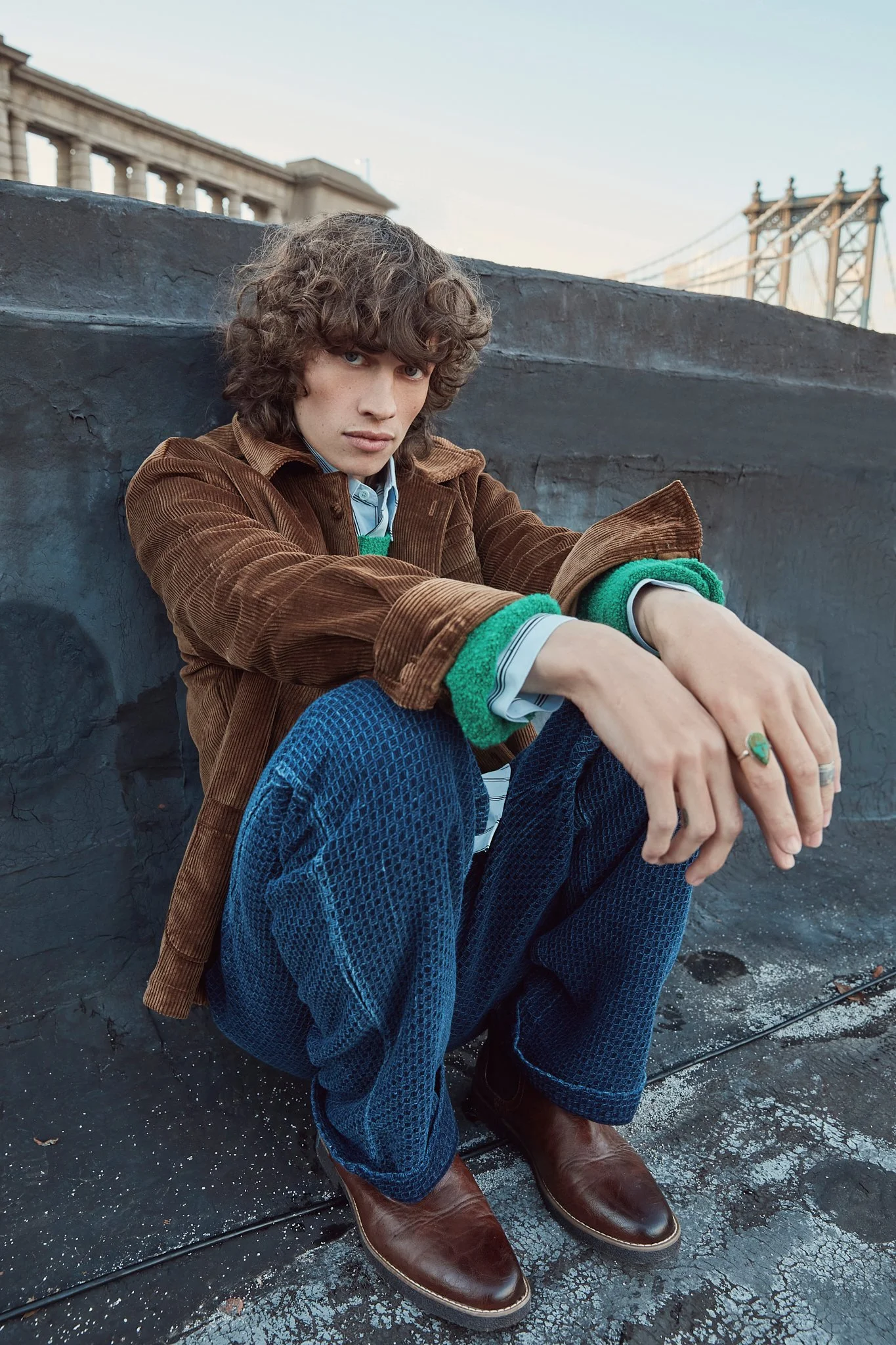 Person with curly hair seated on urban rooftop, wearing a brown corduroy jacket, green sweater, blue textured pants, and brown leather shoes. Cityscape and bridge in the background.