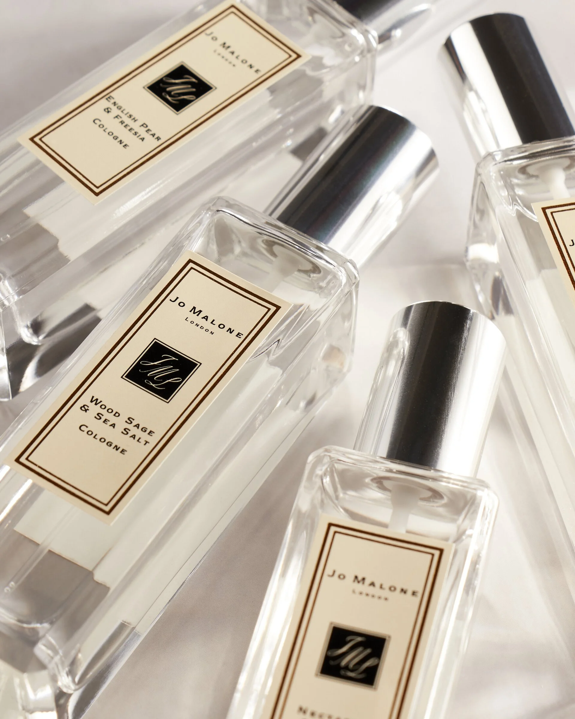 Several Jo Malone cologne bottles with labels including "English Pear & Freesia" and "Wood Sage & Sea Salt," featuring silver caps.