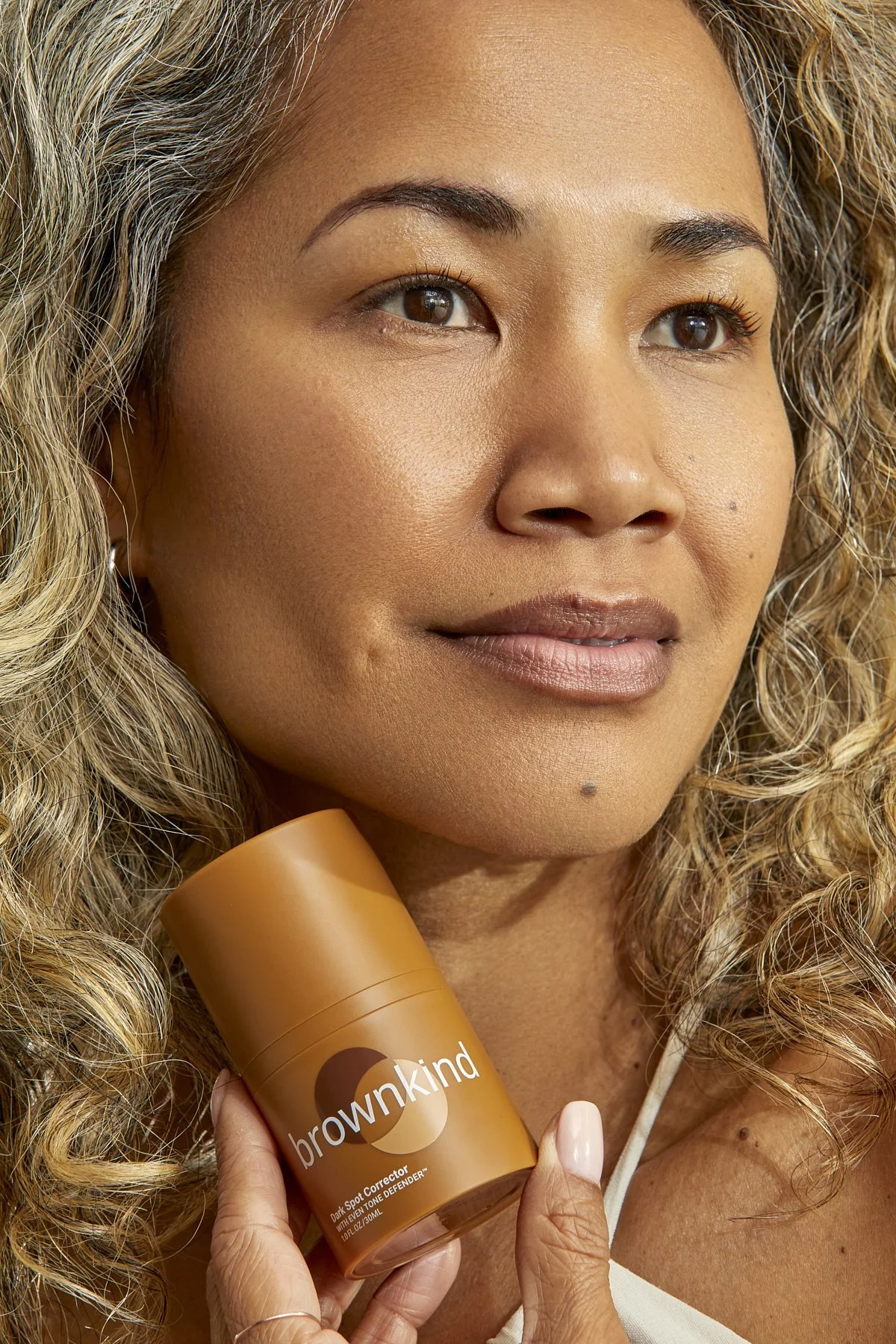 Brownkind skincare campaign: portrait of a woman with curly hair holding the Brownkind Dark Spot Corrector against her cheek, photographed by Adrianna Favero.