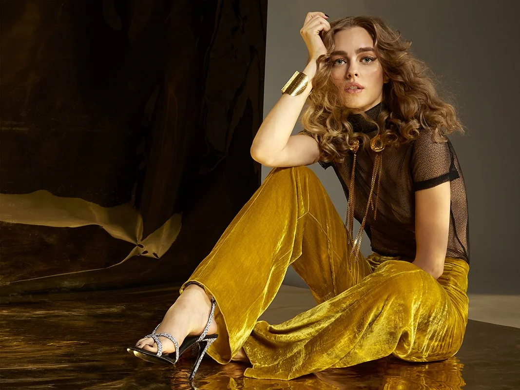 FAJO Magazine holiday fashion editorial: model seated on the floor in mustard wide-leg trousers and a sheer black top, leaning back with hand in her hair, photographed by Adrianna Favero.