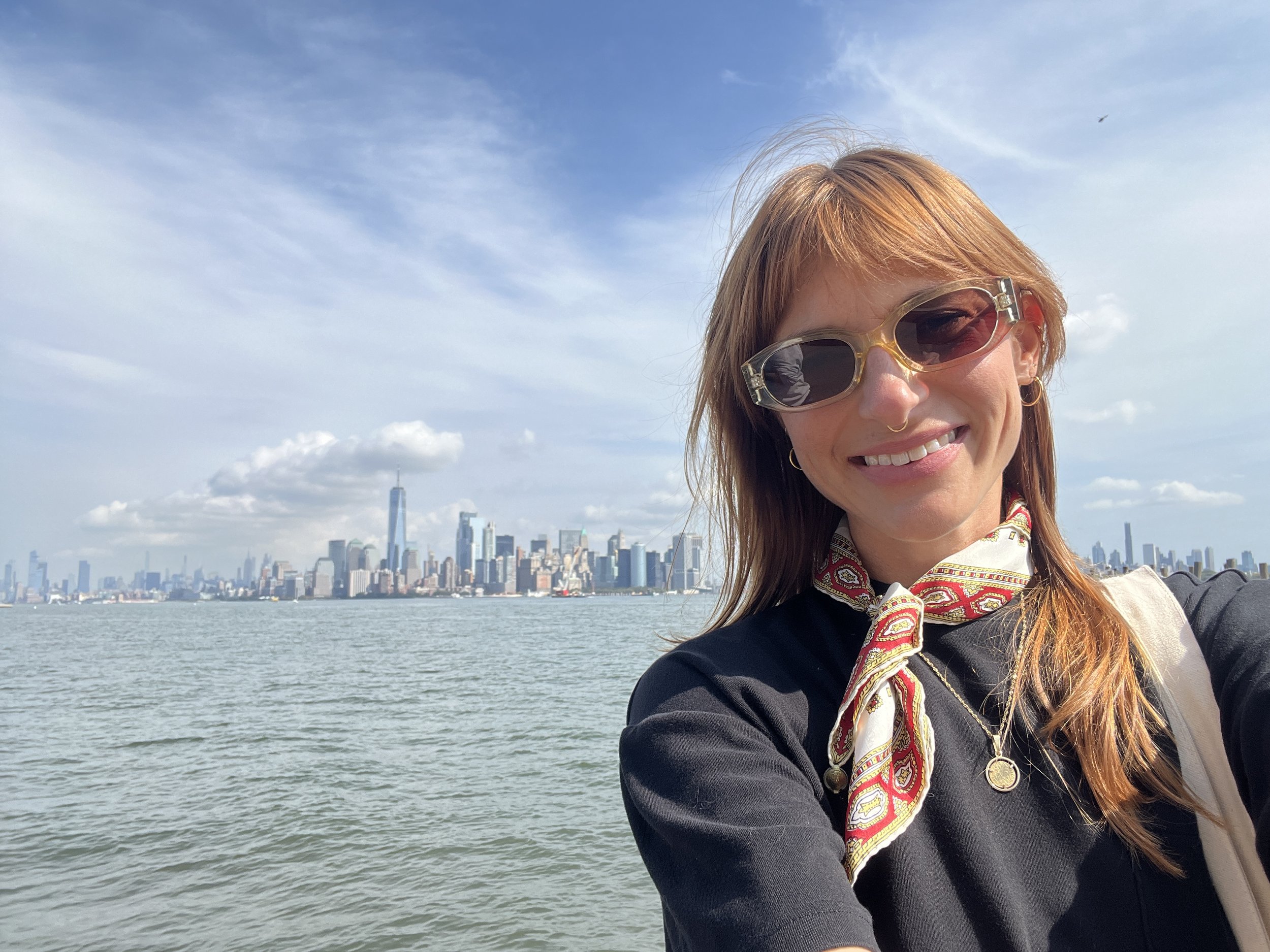 A woman with auburn hair and sunglasses taking a selfie by the water with a city skyline, including One World Trade Center, in the background.