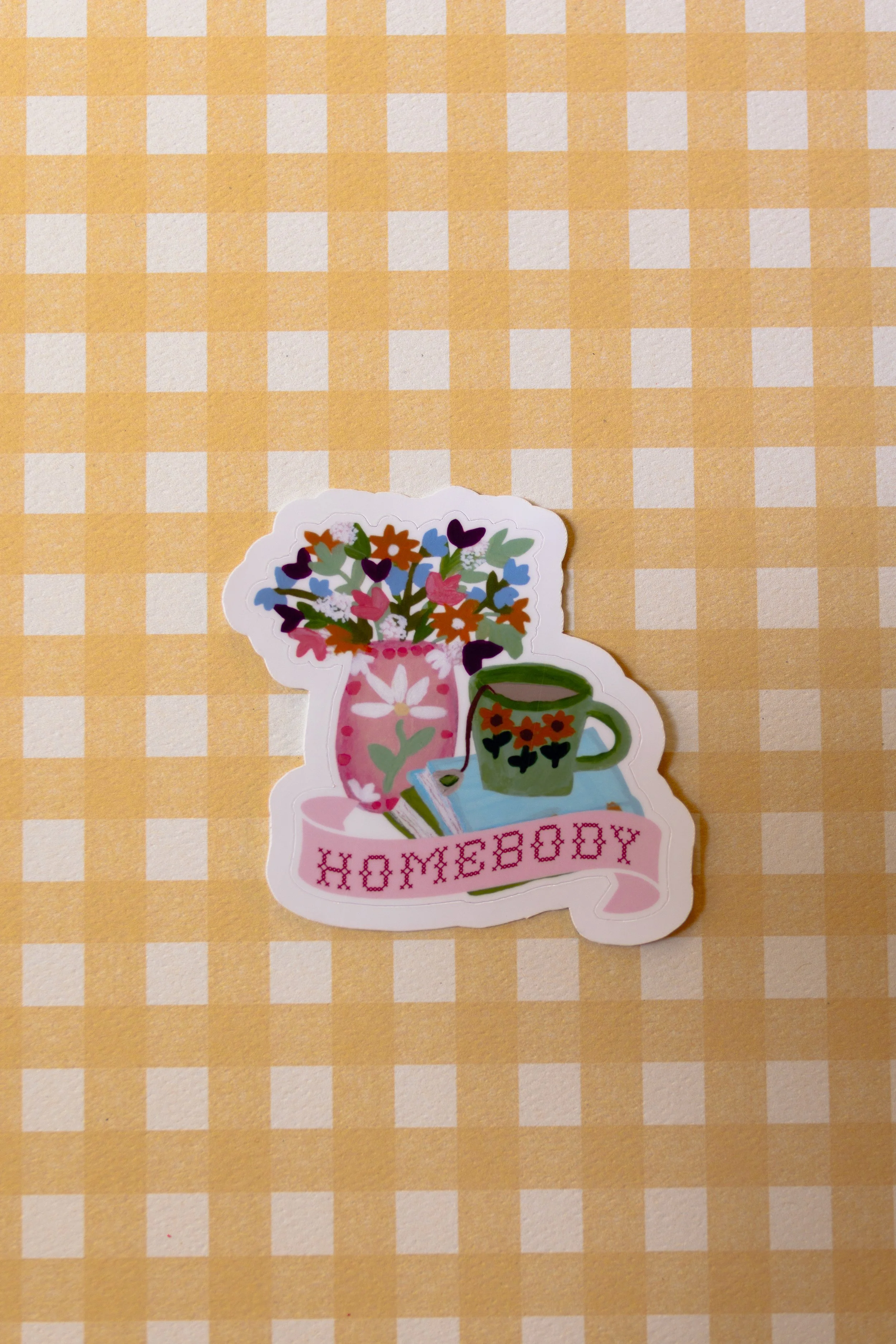 This sticker features a cute vase of flowers, stack of books, and floral teacup that reads “Homebody” in cross stitch letters.