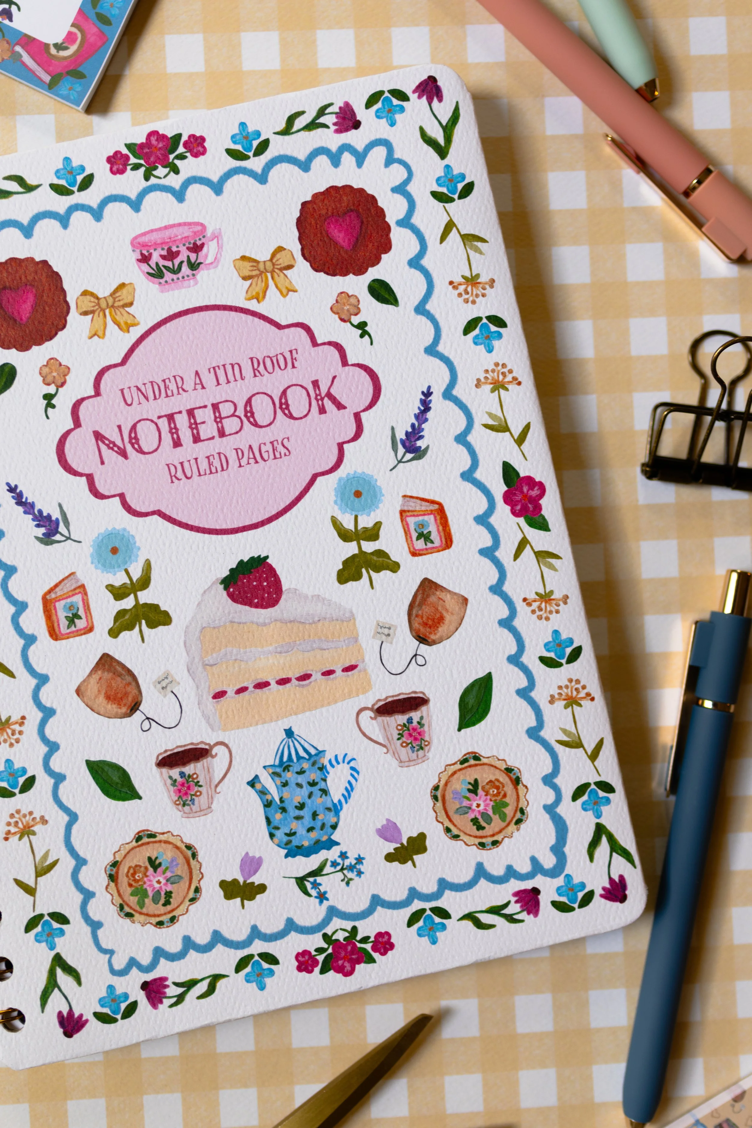 Gold spiral bound ruled notebook with textured cover featuring colorful afternoon tea illustrations.