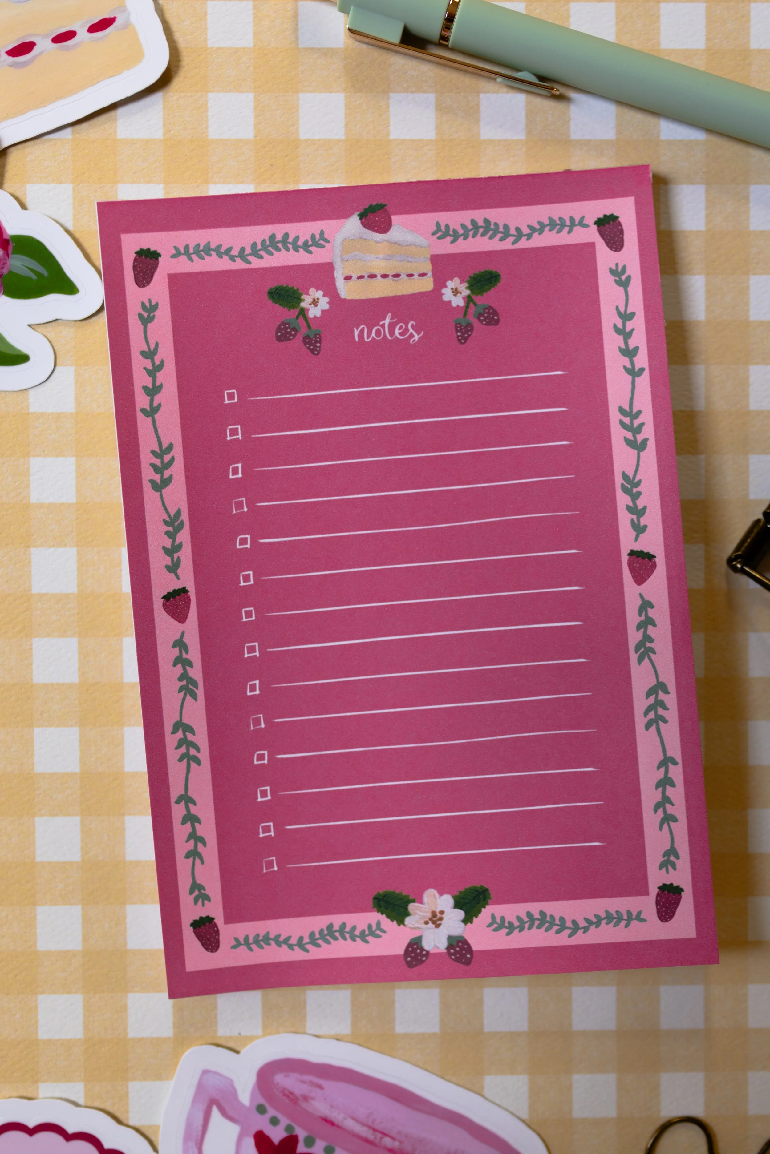 Whimsical strawberry cake notepad displayed beside tea, fresh strawberries, and vintage stationery.