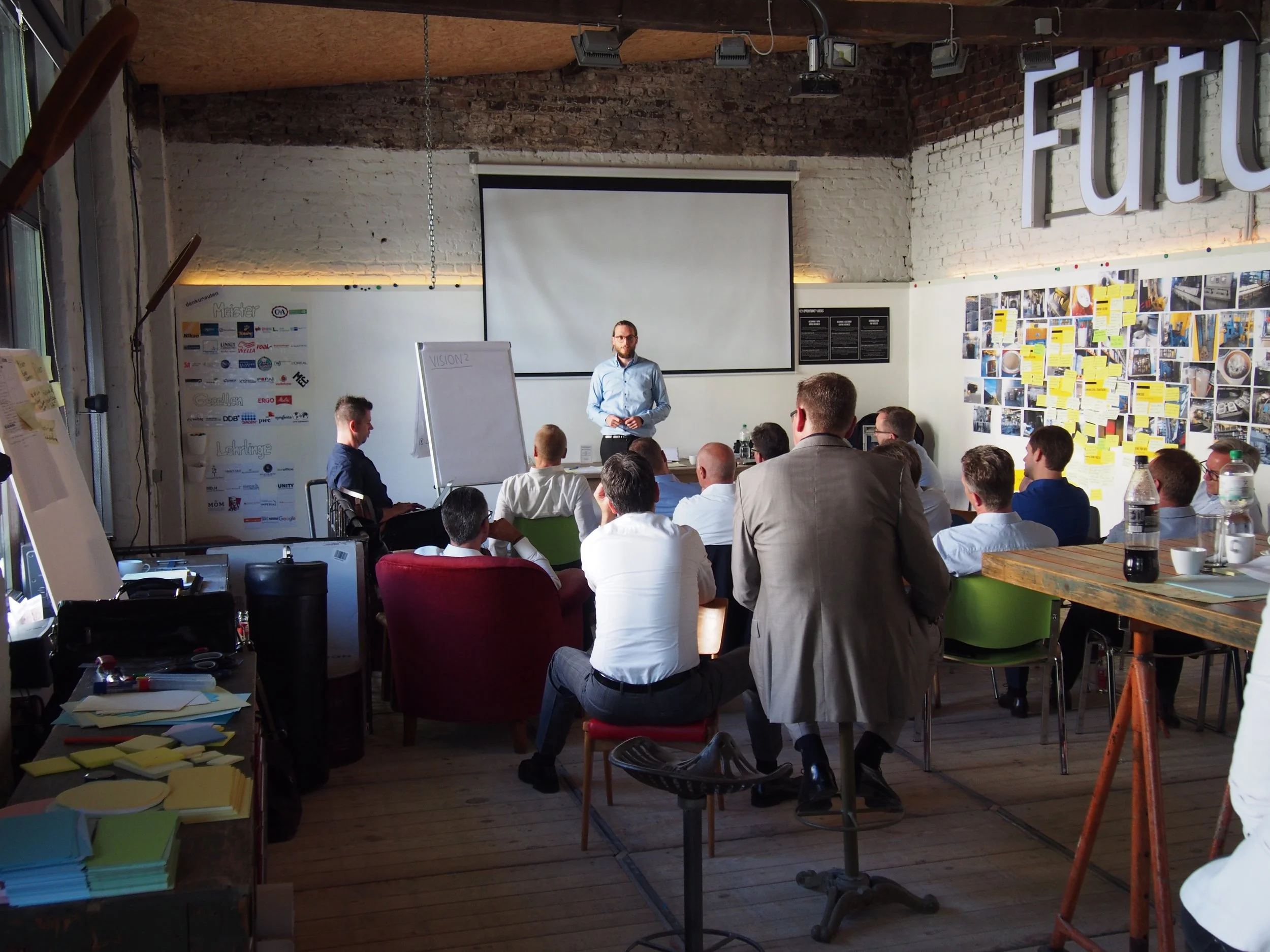 A group of people attending a presentation or meeting in a room with brick walls and large windows. A man is standing in front of a whiteboard, speaking to the audience.