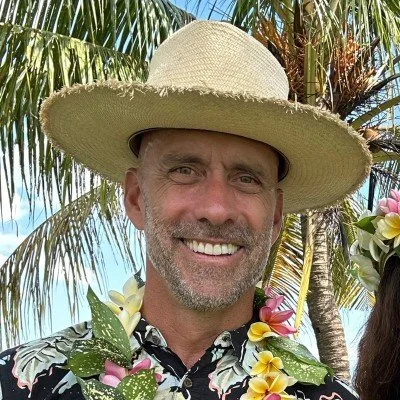 Smiling middle-aged man wearing a large straw hat, floral shirt, tropical flowers, and palm trees in the background on a sunny day.