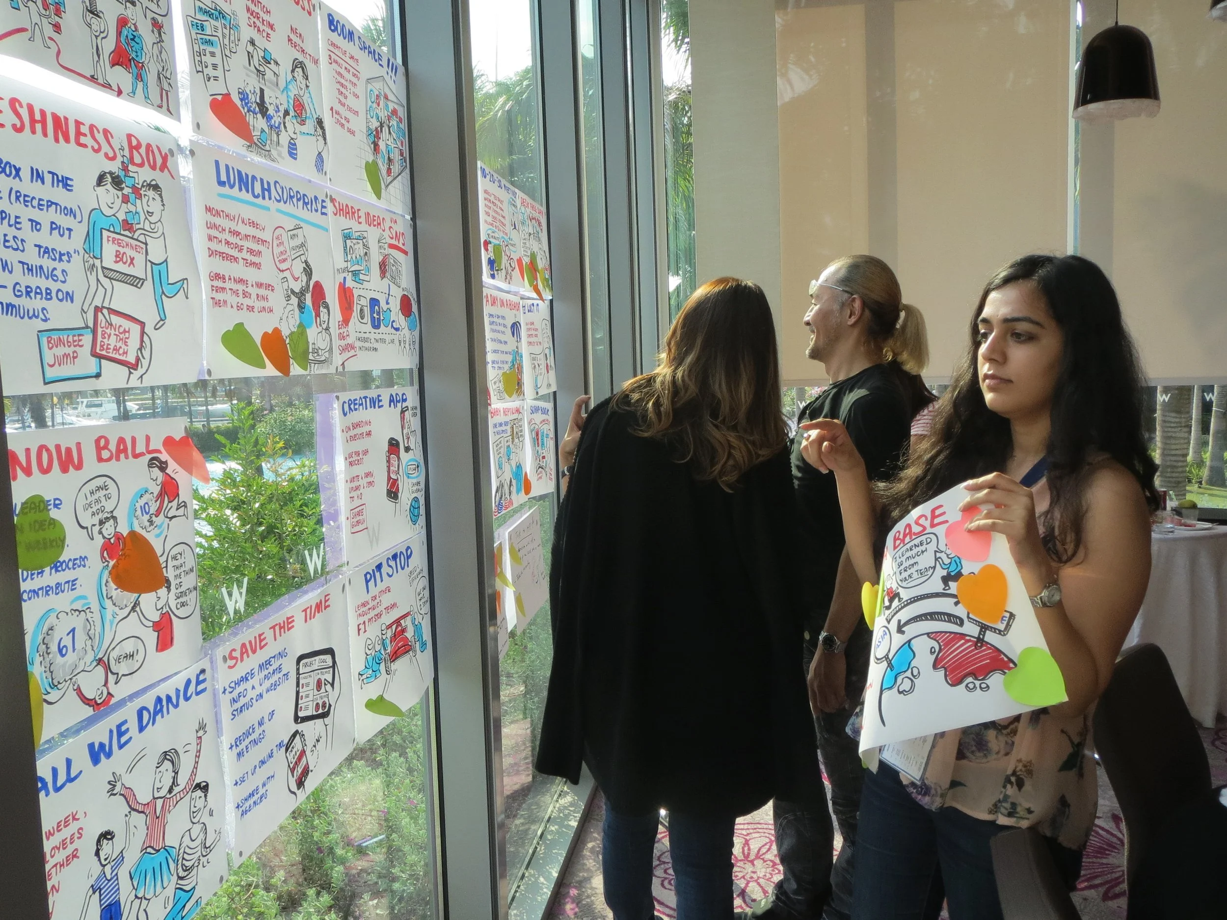 People looking at colorful illustrated posters on a glass wall that contain various advice and tips, with some individuals holding papers or notepads.