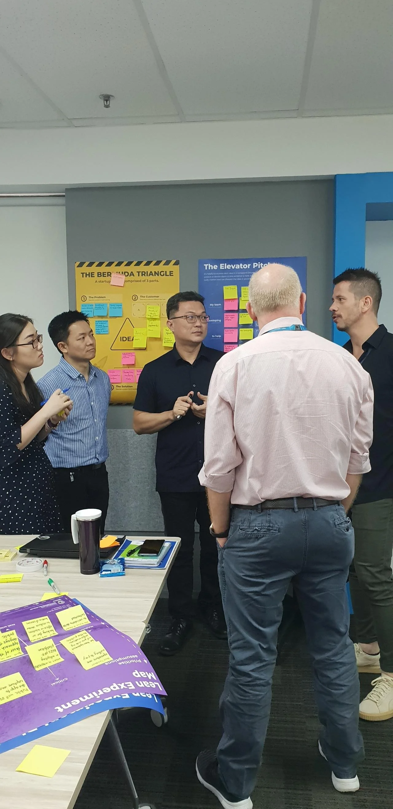 Group of five adults engaged in a discussion in an office or conference room, with posters, sticky notes, and office supplies on the table and walls.