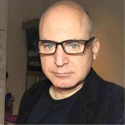 A man with light skin, glasses, and a shaved head wearing a black shirt and blazer, taking a selfie indoors.