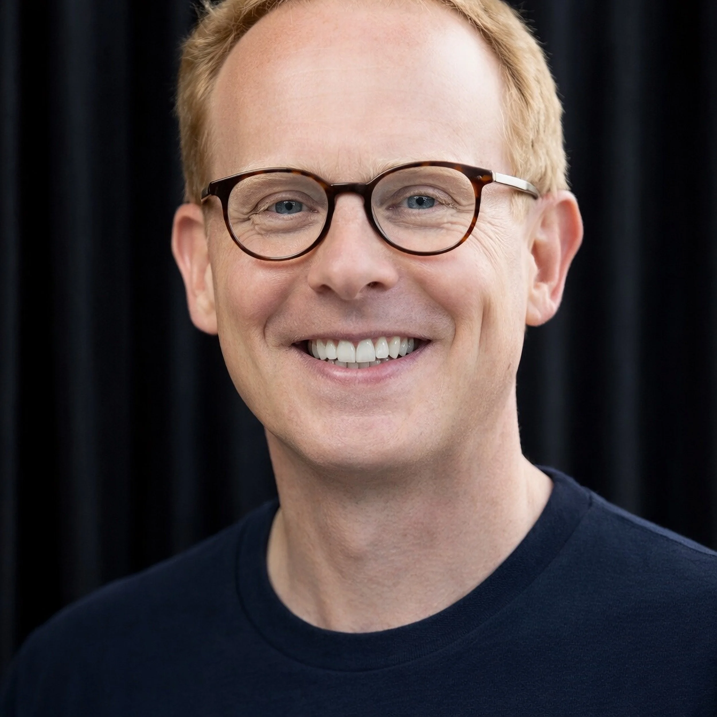 Portrait of Josh Woodward, a senior Google product leader, smiling and wearing round glasses against a dark, softly blurred background.
