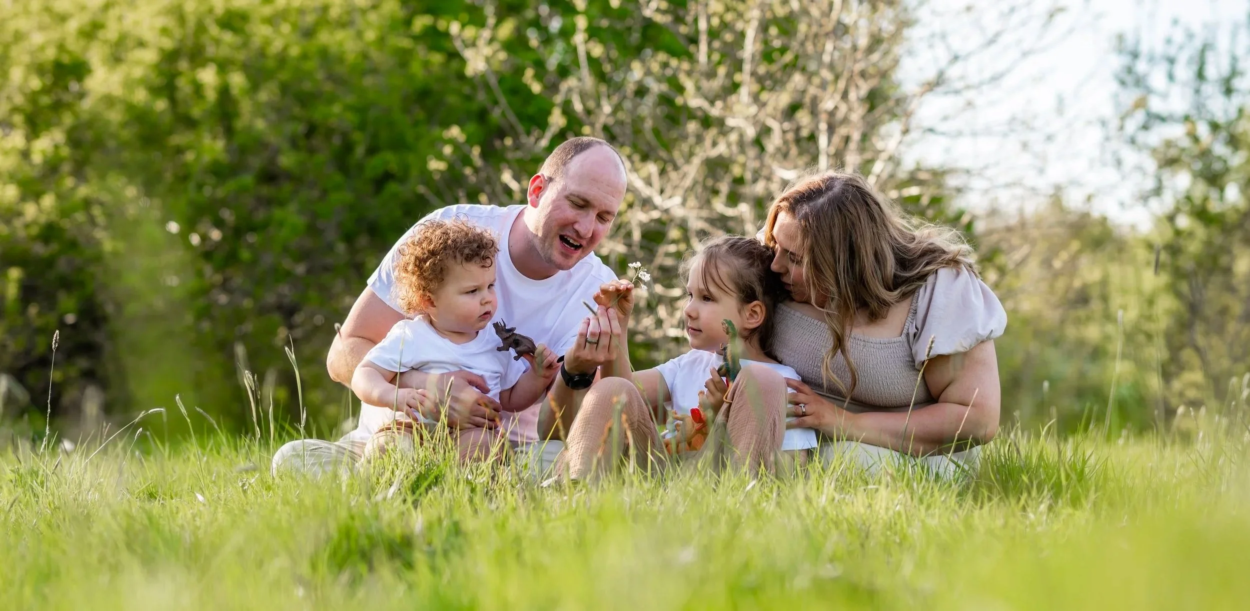 Mum, Dad and two young boys sit barefoot in a field at sunset, they're looking at flowers dad is holding, smiling. They are wearing beige and white.