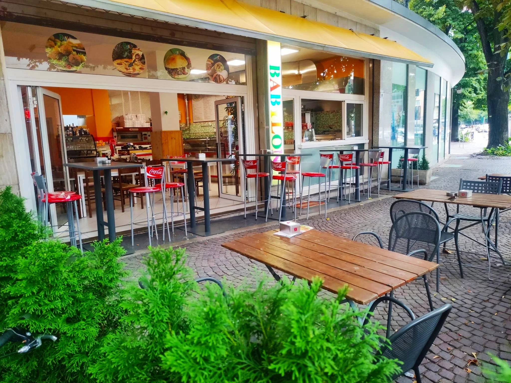 Esterno di un ristorante con terrazza, tavoli di legno, sedie metalliche e un'insegna luminosa. Il locale ha tendalino giallo e decorazioni interne. "BABILON" è scritto sull'insegna."