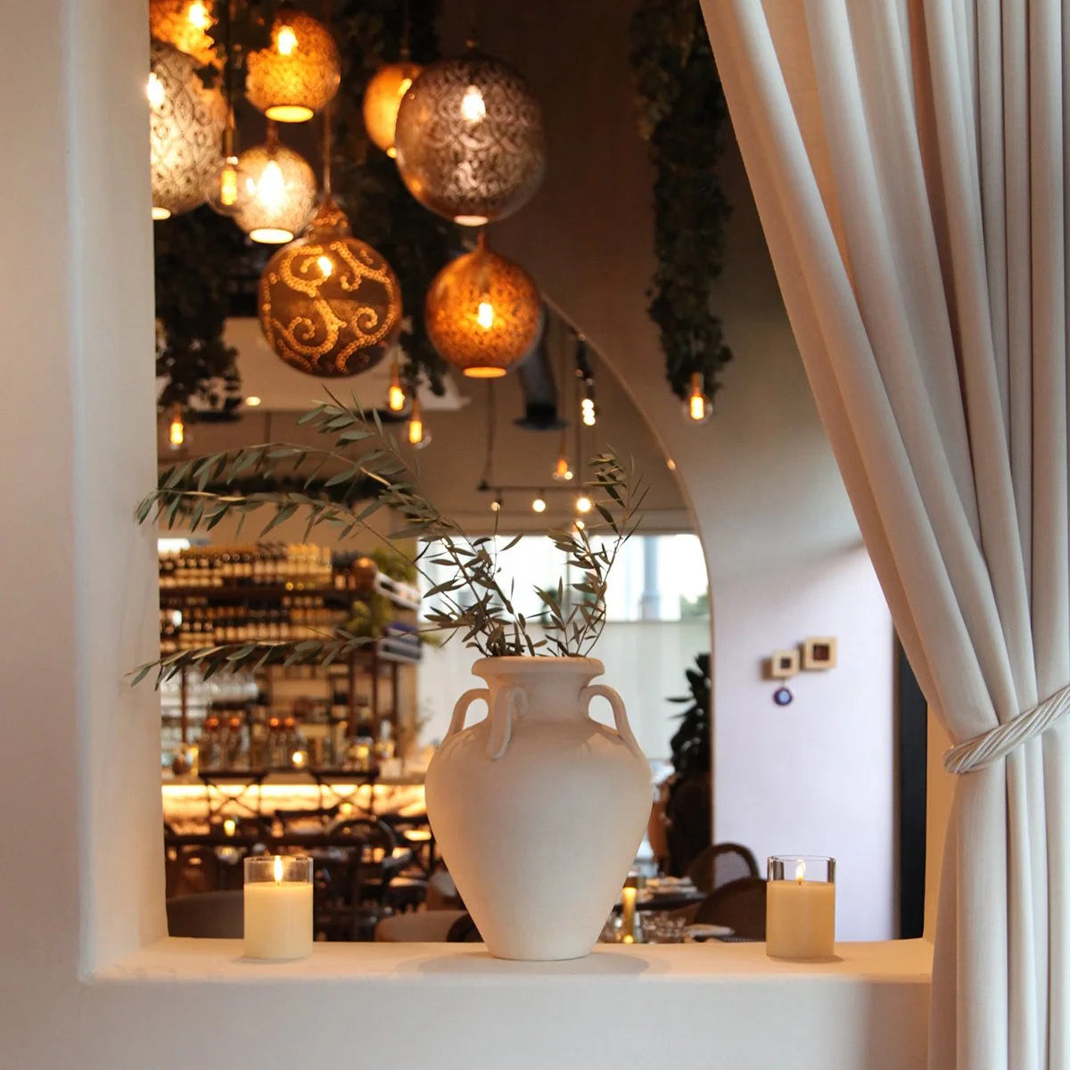 alt="Evening shot of the Ammatoli dining room with warm golden light tones, an ivory curtain, hanging brass lanterns, candles, antique-style pottery with fresh-cut olive branches displayed""