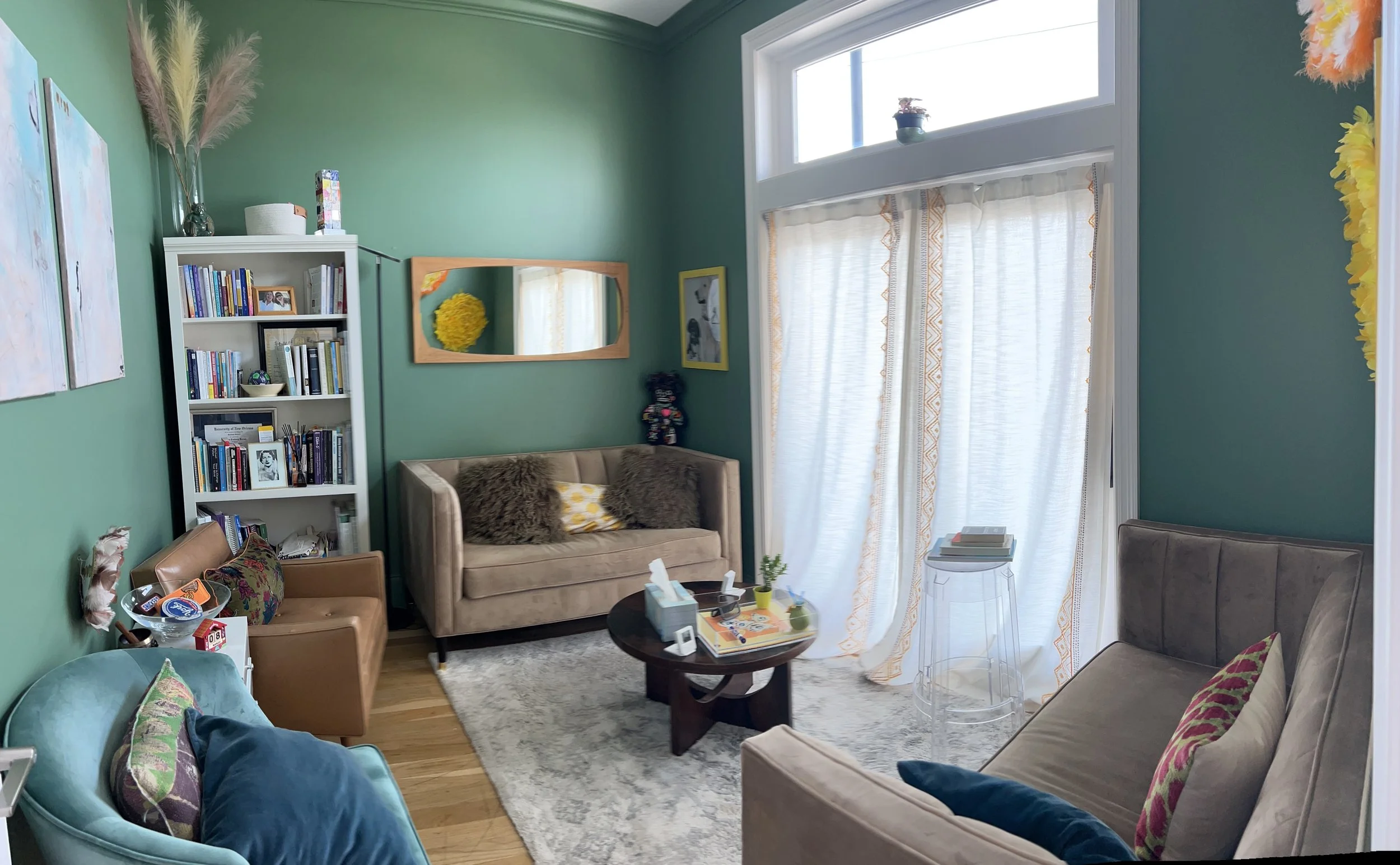 A cozy living room with light green walls and large windows with white curtains. The room features a beige armchair with fuzzy pillows, a light blue armchair with patterned pillows, a dark wood coffee table with books and decorative items, a white bookshelf filled with books and framed photos, and a mirror on the wall. There are various decorative accents including a vase with pampas grass, colorful artwork, and a small side table with tissues and plants.