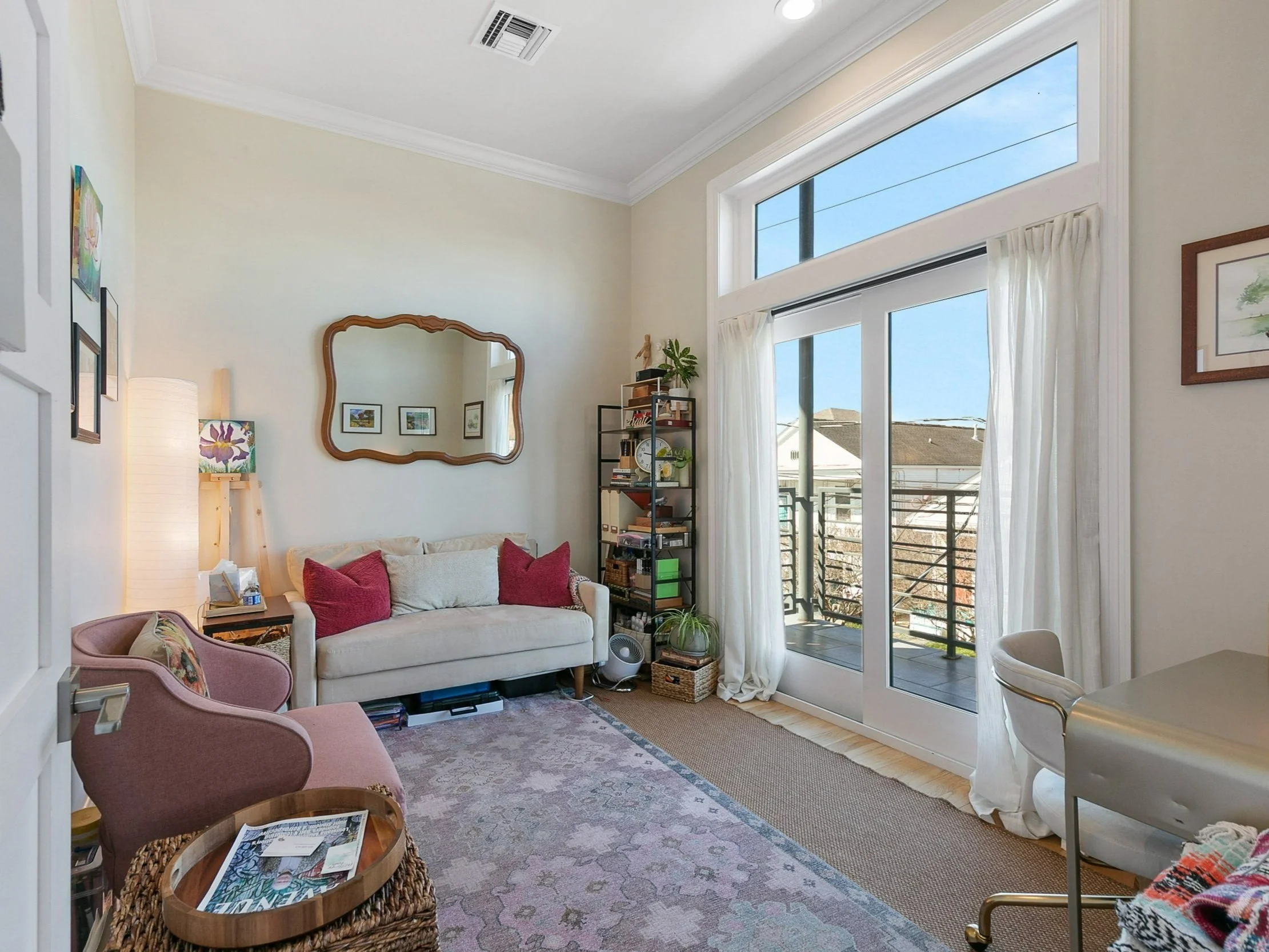 Living room with large windows and curtains, beige and pink furniture, artwork on the walls, bookshelf with decorations, and a glass sliding door leading to a balcony.