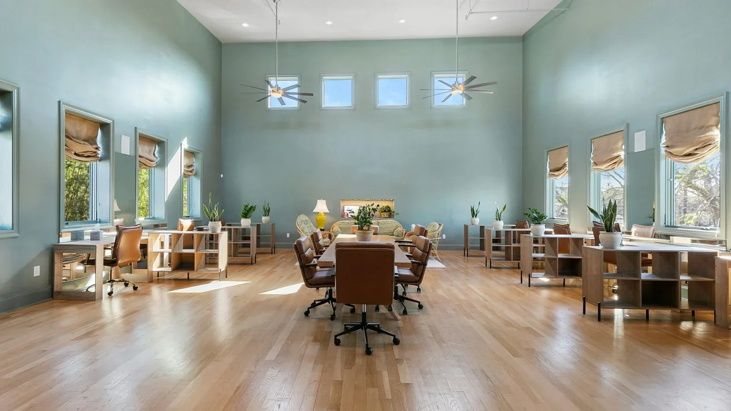 A spacious conference room with high ceilings, green walls, multiple windows with beige shades, and wooden flooring. The room has a large rectangular table with brown chairs, potted plants, and additional seating near the back wall, which features a sofa, chairs, and a lamp.