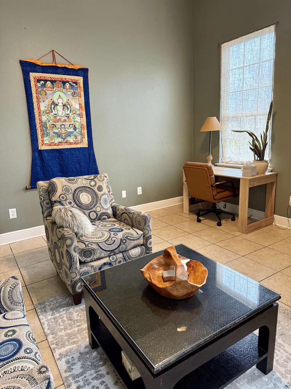 Lobby of private office space rental building with stone tile floors, a square black stone coffee table, a wooden desk and leather desk chair, a gray rug, an arm chair with blue and gray circles, and a textile of an Indian diety on the wall
