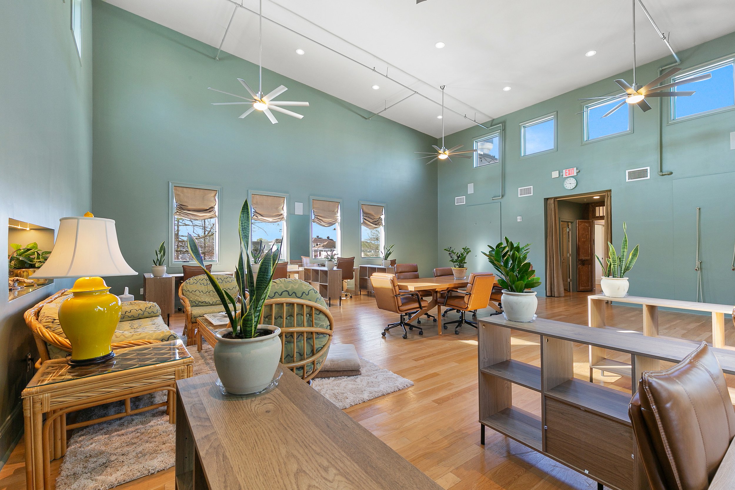 A spacious, well-lit indoor lounge or common area with high ceilings, multiple windows, ceiling fans, and various seating arrangements including chairs, couches, and tables. The room is decorated with potted plants and has light-colored hardwood floors.